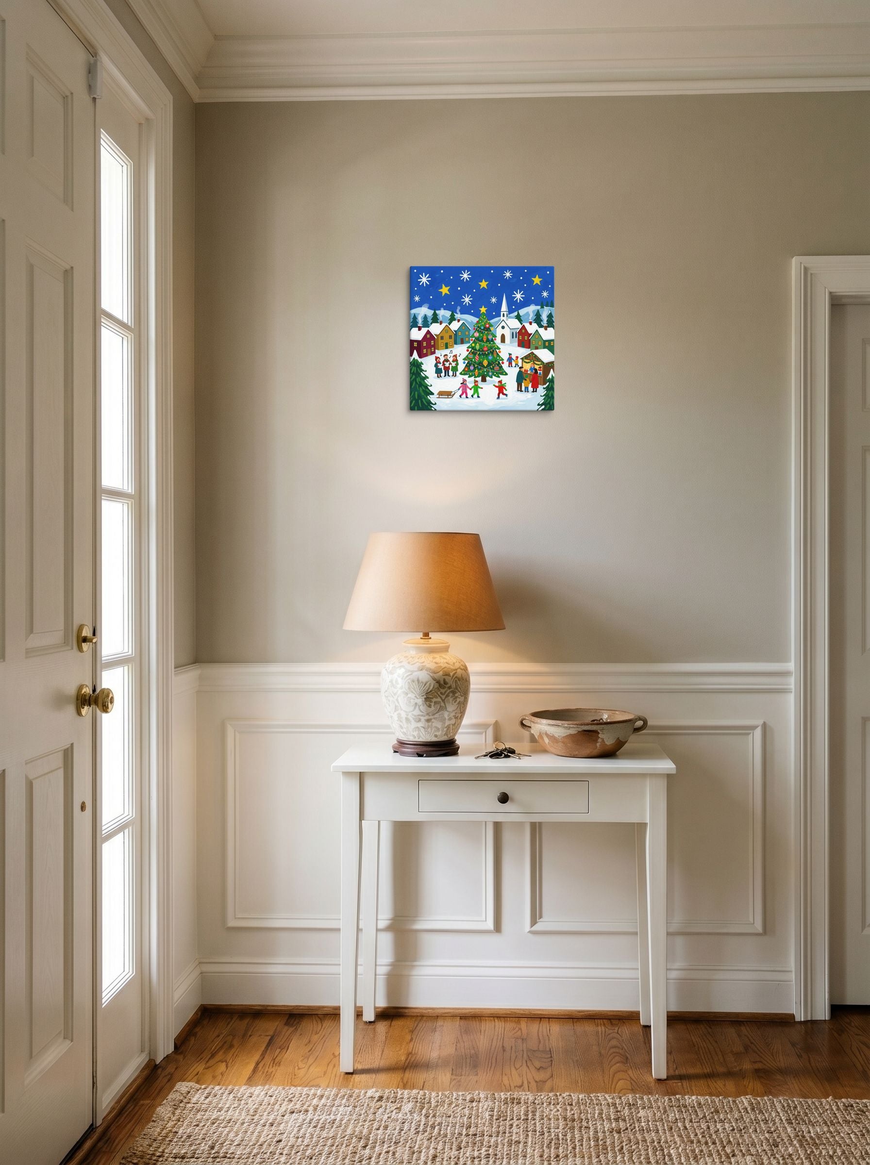 A 12x12 inch canvas Naive art print of 'Joyous Christmas Village' hanging in an entryway near a console.