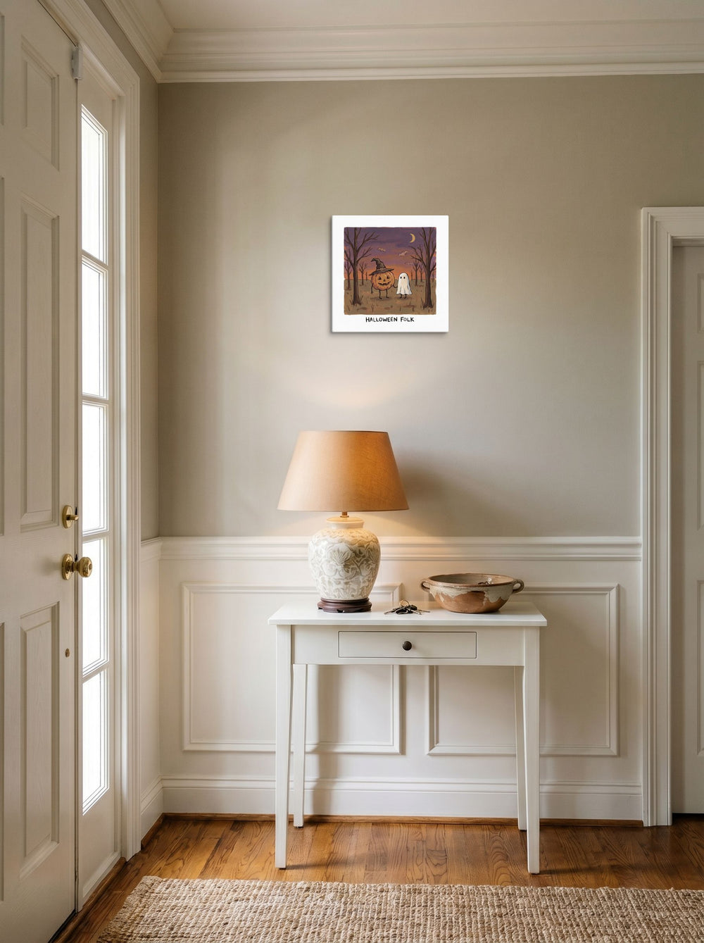 A 12x12 inch canvas Naive art print of 'Halloween Folk' hanging in an entryway near a console.
