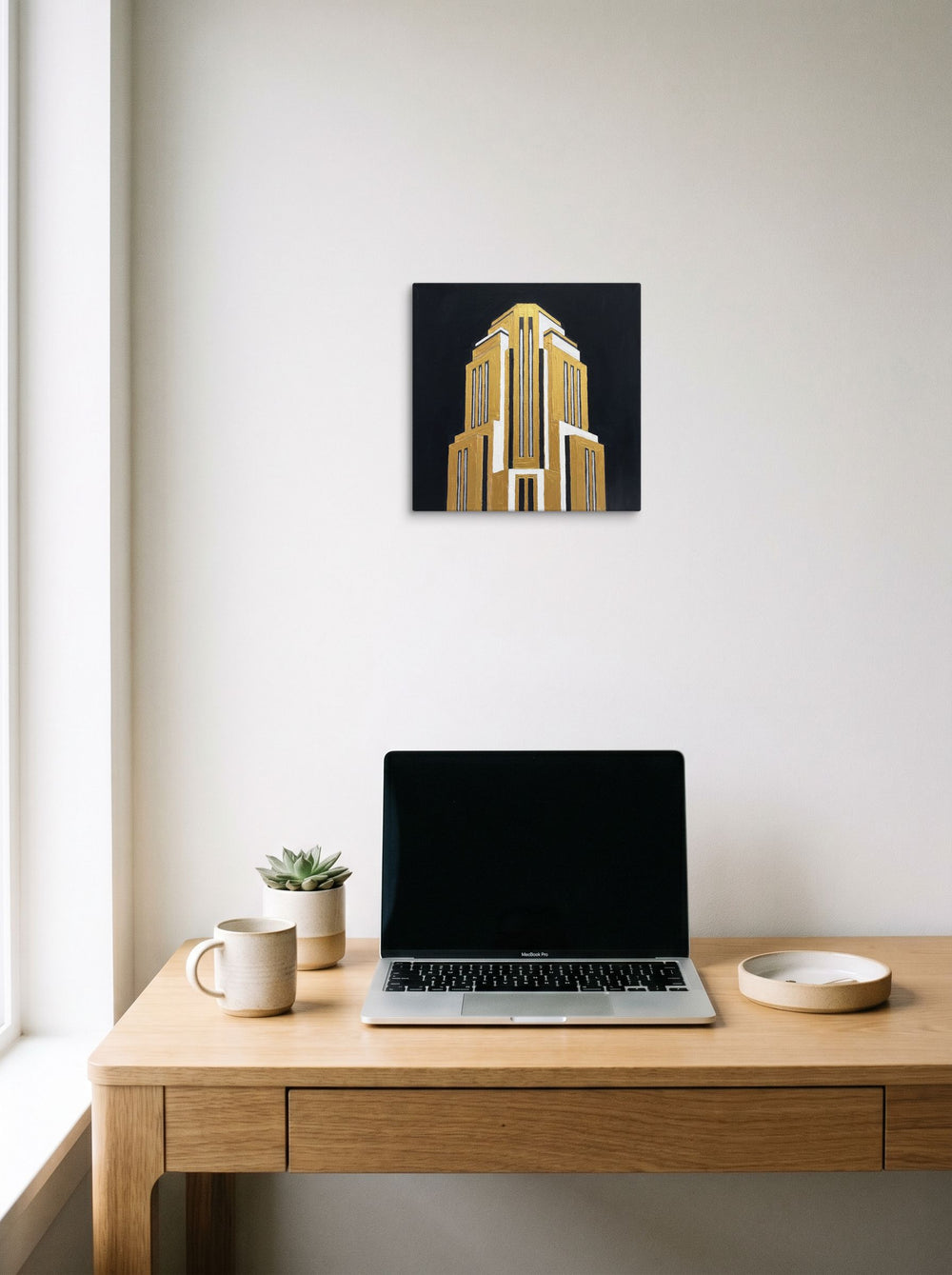 A 12x12 inch canvas Art Deco print of 'Tower of the Jazz Age' hanging above a worktable with a laptop.