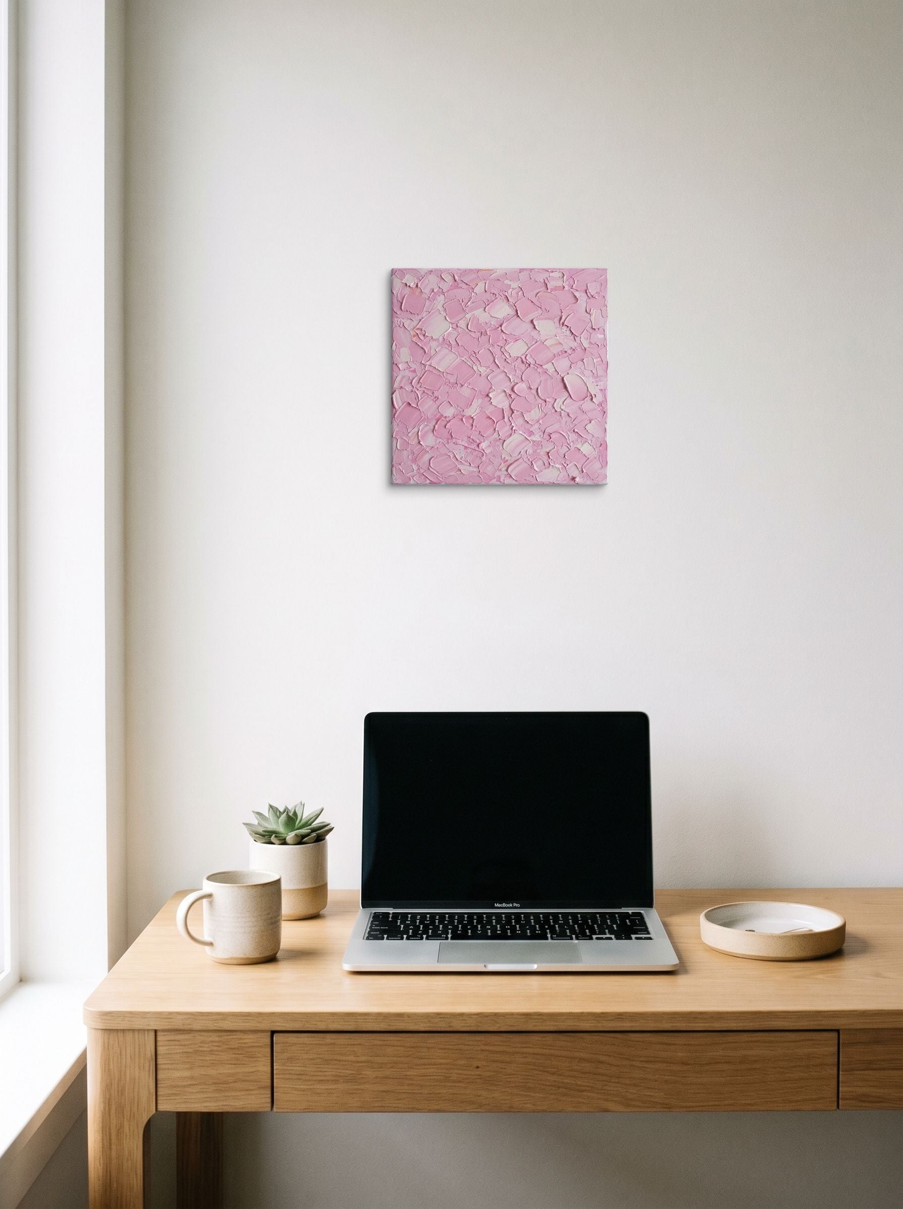 A 12x12 inch canvas Expressionism print of 'Blush Topography' hanging above a worktable with a laptop.