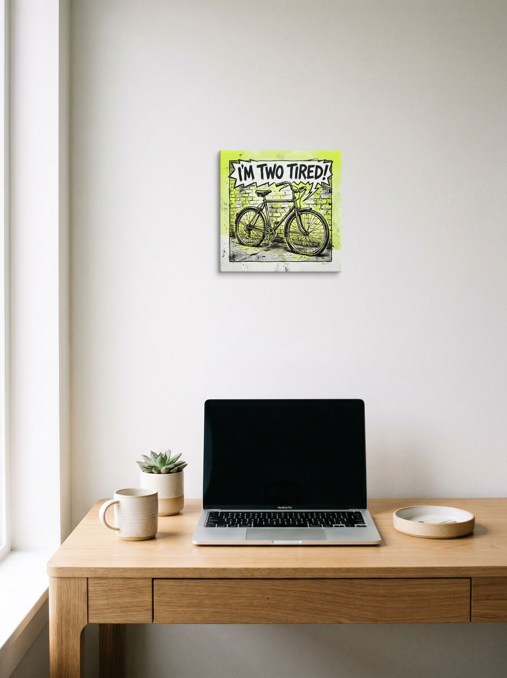 A 12x12 inch canvas Pop art print of 'Two Tired' hanging above a worktable with a laptop.
