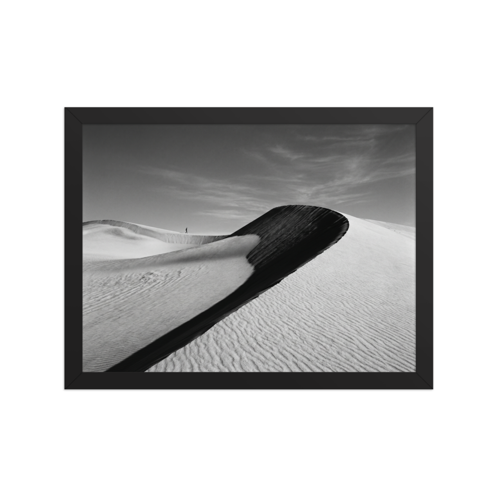 'Footsteps in the Void' — a Art photography print. A minimalist black and white photograph of sweeping sand dunes with a tiny figure walking along the ridge, perfect for adding a sense of scale to an Office.