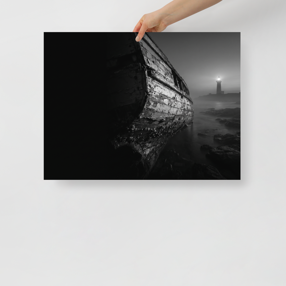 'Final Anchor' — a Art photography print. Black and white photograph of an old, peeling wooden boat resting on rocks with a glowing lighthouse in the foggy distance, perfect for a cozy living room.