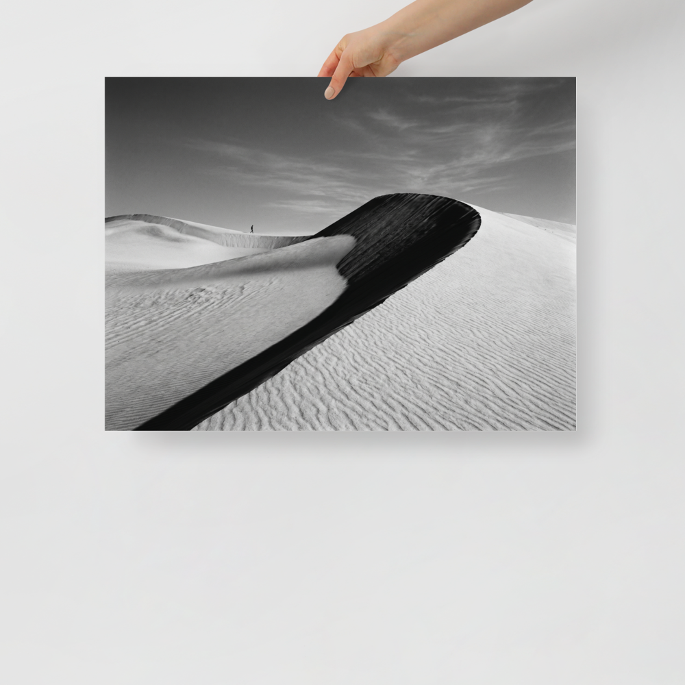 'Footsteps in the Void' — a Art photography print. A minimalist black and white photograph of sweeping sand dunes with a tiny figure walking along the ridge, perfect for adding a sense of scale to an Office.