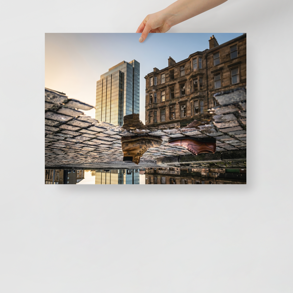 'Steps Through Time' — a Art photography print. Creative puddle reflection photograph showing a work boot and a dress shoe on cobblestones, with a modern glass skyscraper and an old stone building in the background, ideal for an office or hallway.