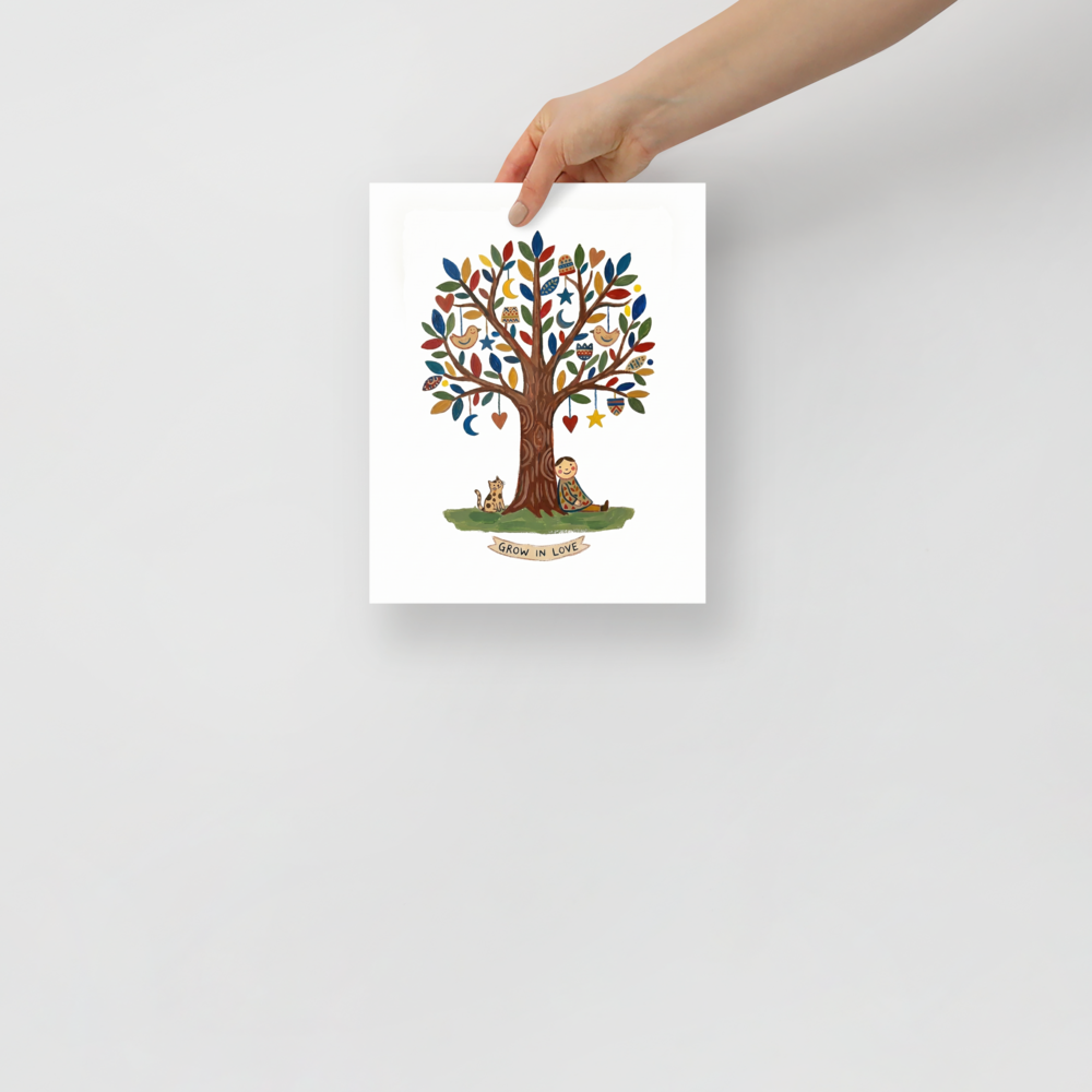 'The Tree Of Love' — a Naive art print. A heartwarming naive art piece showing a person and a spotted cat sitting under a tree decorated with hearts and moons, with a banner reading GROW IN LOVE.