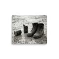 'The Drifter's Boots' — a American realism print. A black and white photograph of old, worn leather boots resting on cracked earth next to tin cans, perfect for a rustic Living Room.