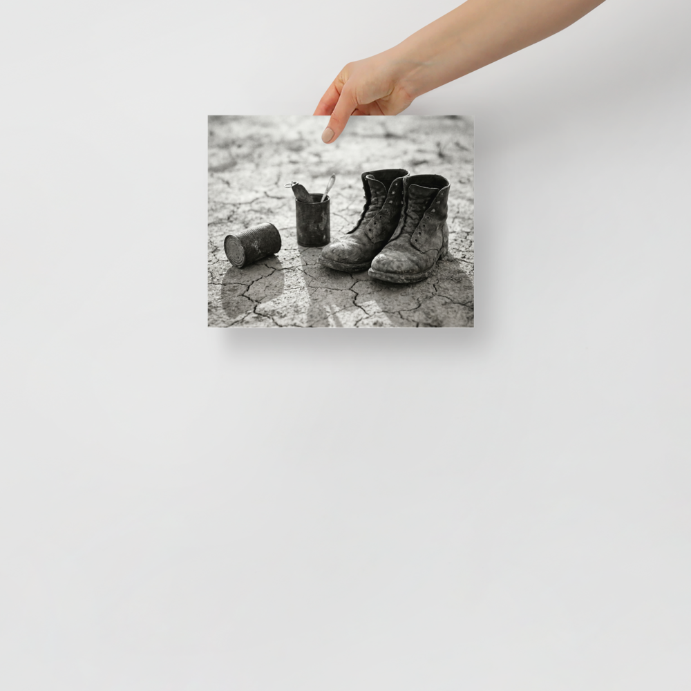 'The Drifter's Boots' — a American realism print. A black and white photograph of old, worn leather boots resting on cracked earth next to tin cans, perfect for a rustic Living Room.