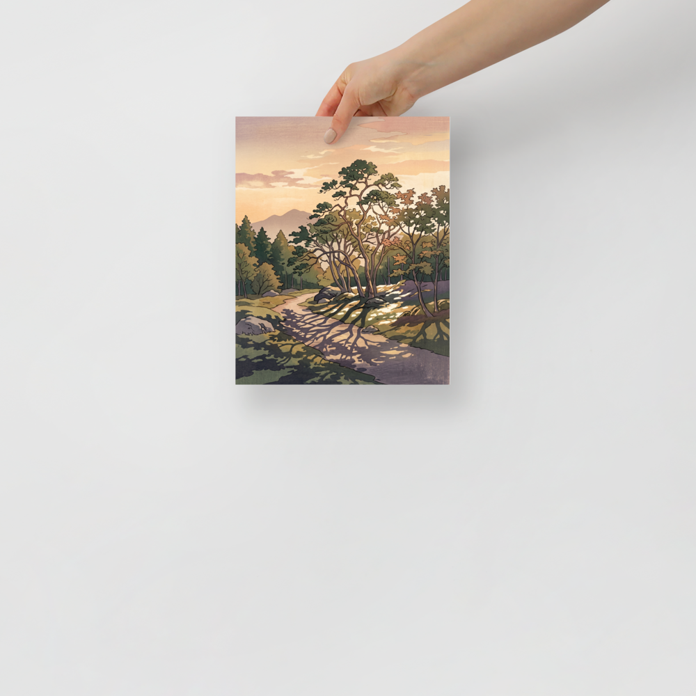 A winding forest path dappled with tree shadows leads toward distant mountains under a warm peach evening sky, ideal for a tranquil living room. A calmness piece titled 'Dappled Sunset Path'.