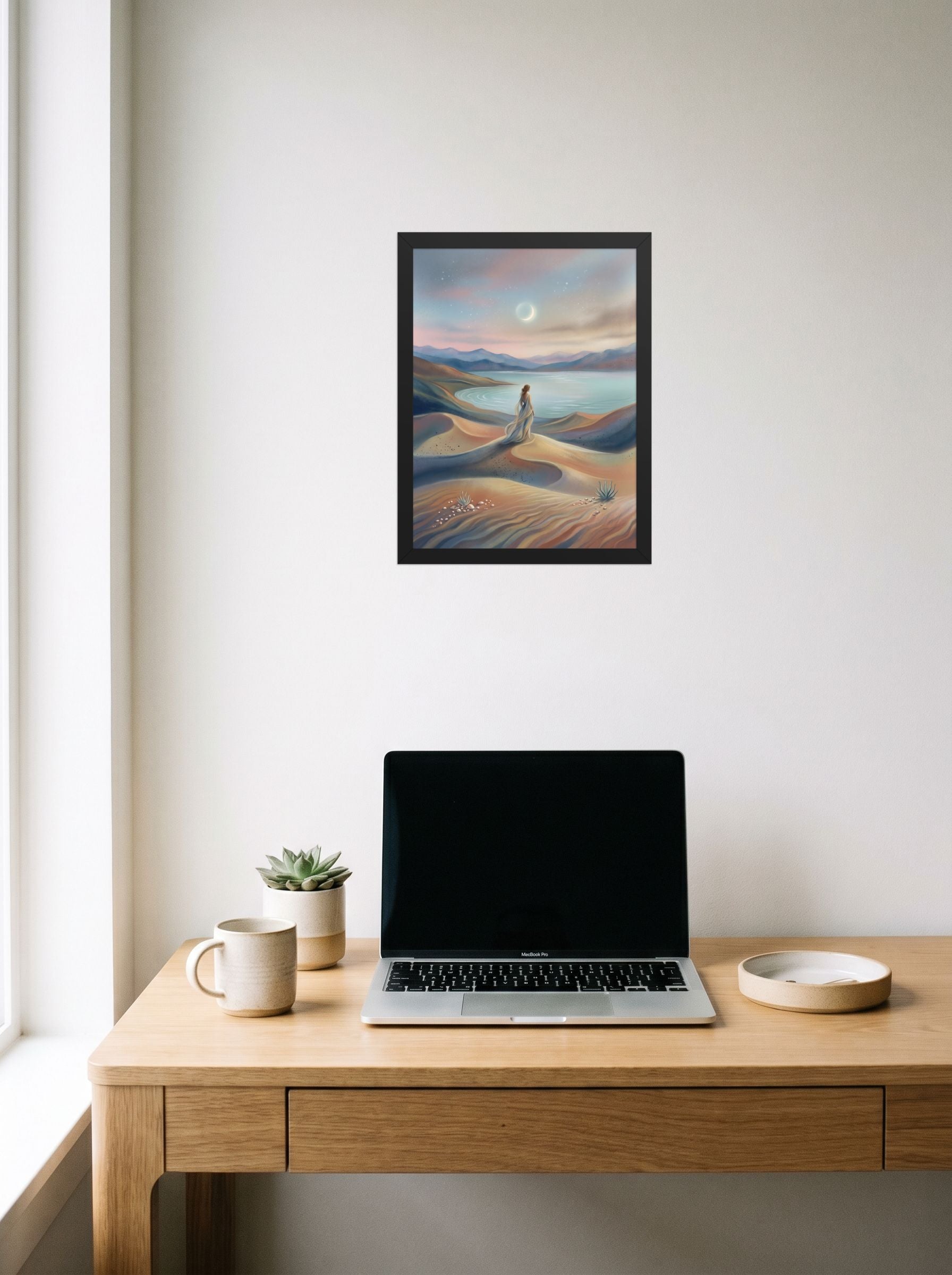 A 12x16 inch black framed Magic realism print of 'Desert Moonrise' hanging above a worktable with a laptop.