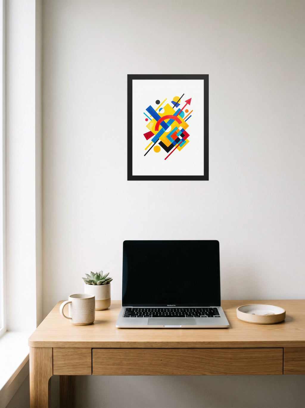 A 12x16 inch black framed Suprematism print of 'Directional Energy' hanging above a worktable with a laptop.