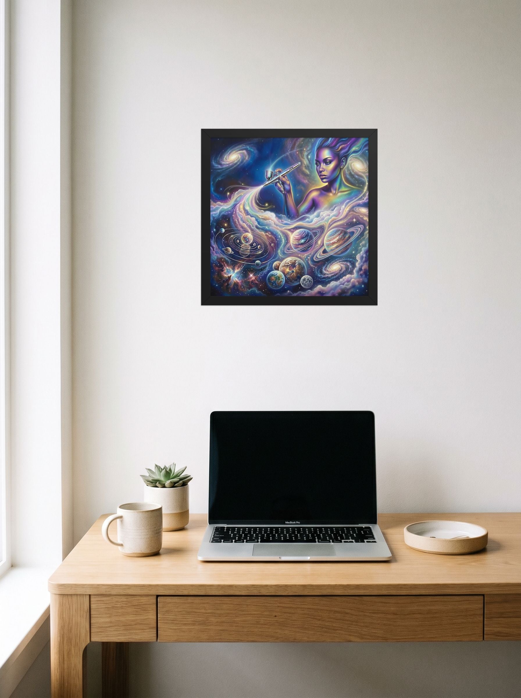 A 14x14 inch black framed Space art print of 'The Cosmic Painter' hanging above a worktable with a laptop.