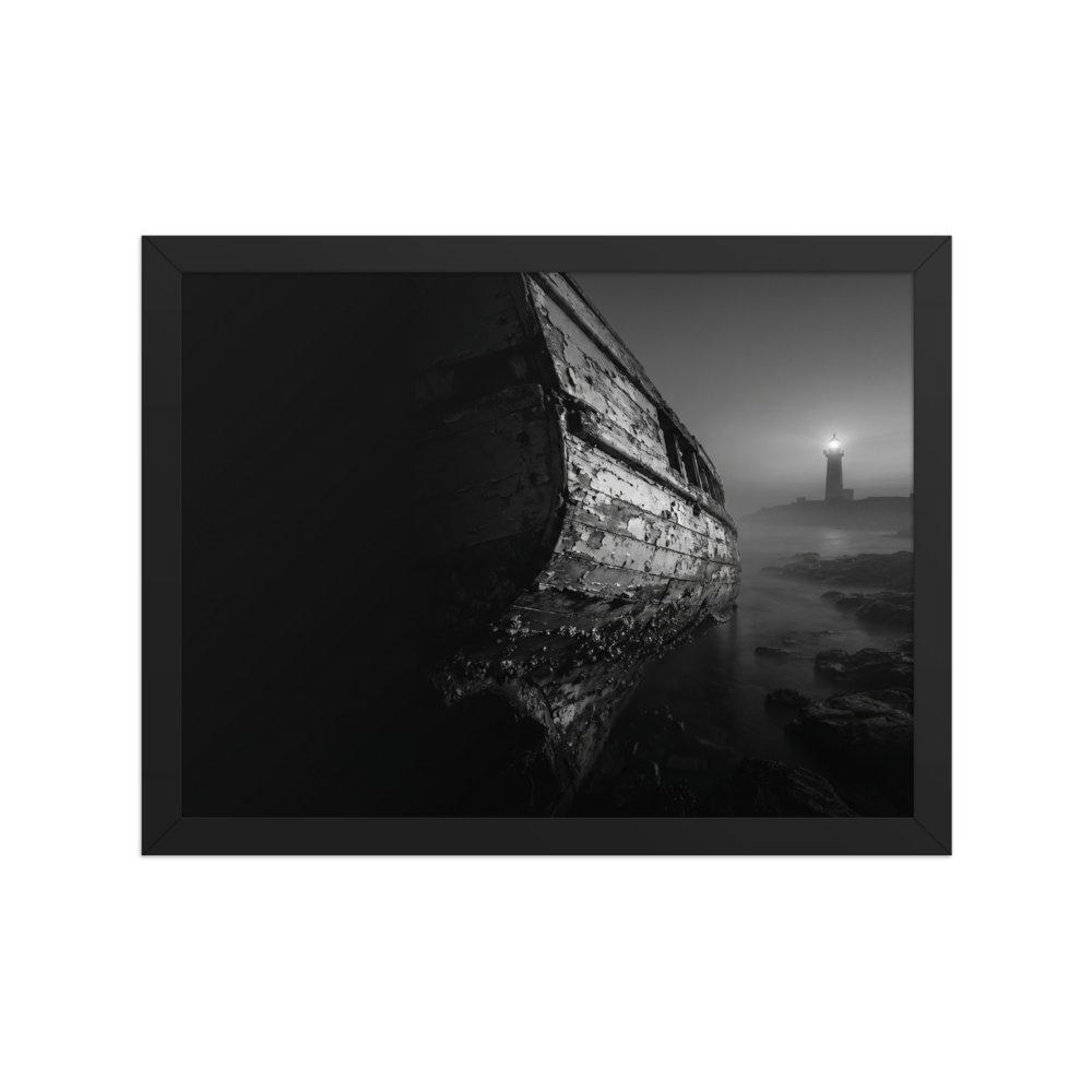 'Final Anchor' — a Art photography print. Black and white photograph of an old, peeling wooden boat resting on rocks with a glowing lighthouse in the foggy distance, perfect for a cozy living room.