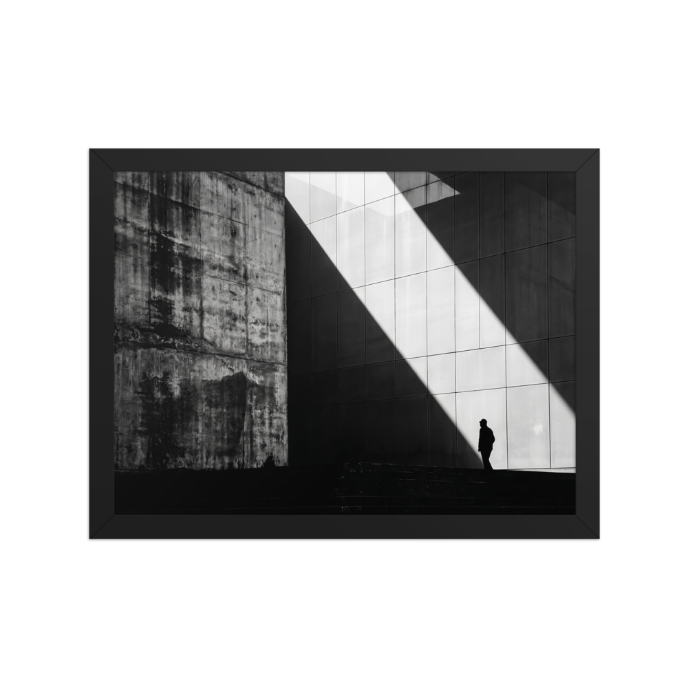 'Urban Geometry' — a Modernism print. A high-contrast black and white architectural photograph featuring a sharp diagonal shaft of light and a lone silhouette walking past textured concrete walls, perfect for an Office or Modern Living Room.