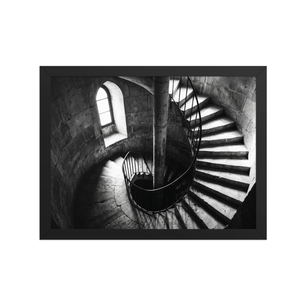 'Descent into Light' — a Art photography print. A high-contrast black and white photograph looking down a spiral stone staircase illuminated by a small window, adding depth and intrigue to an Office or Hallway.