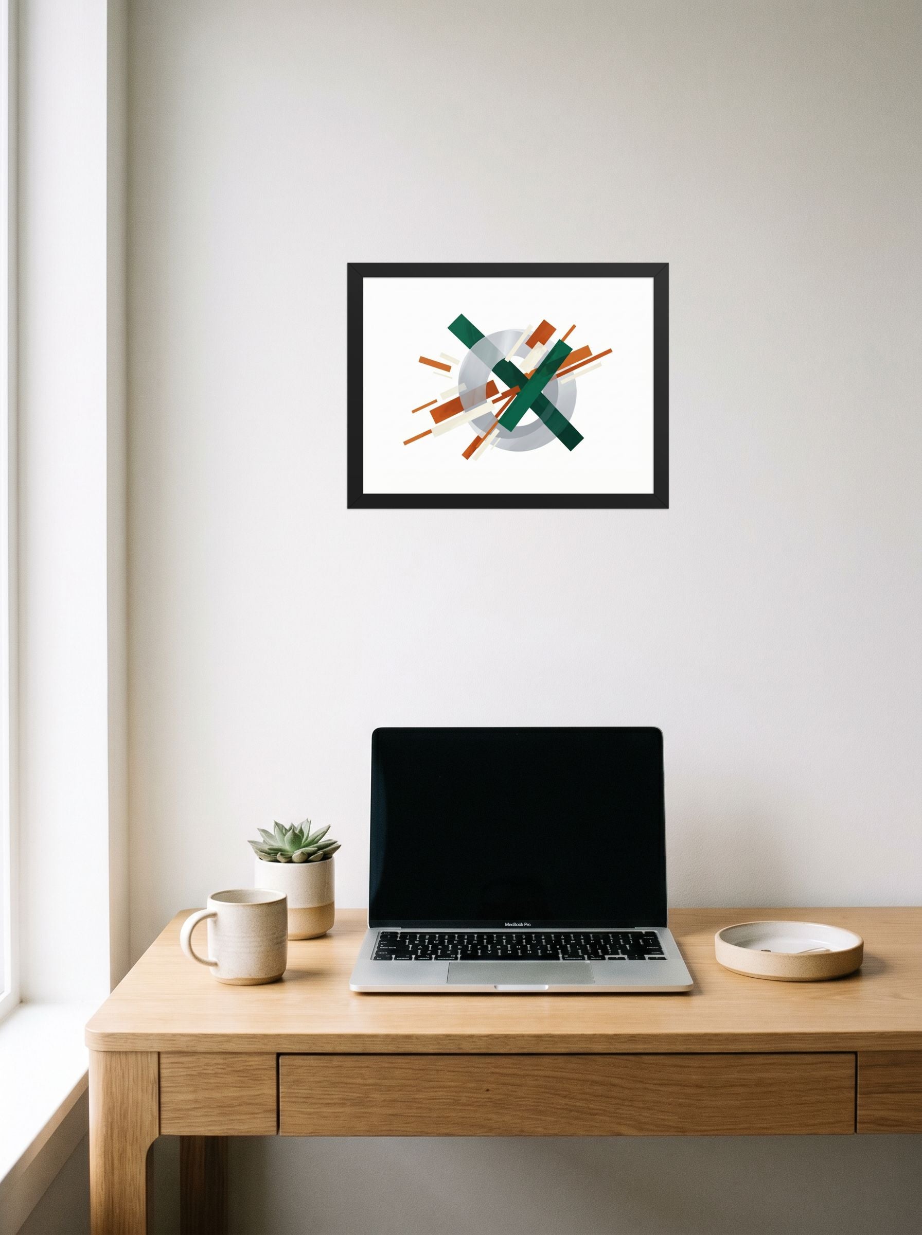 A 16x12 inch black framed Suprematism print of 'Intersecting Perspectives' hanging above a worktable with a laptop.