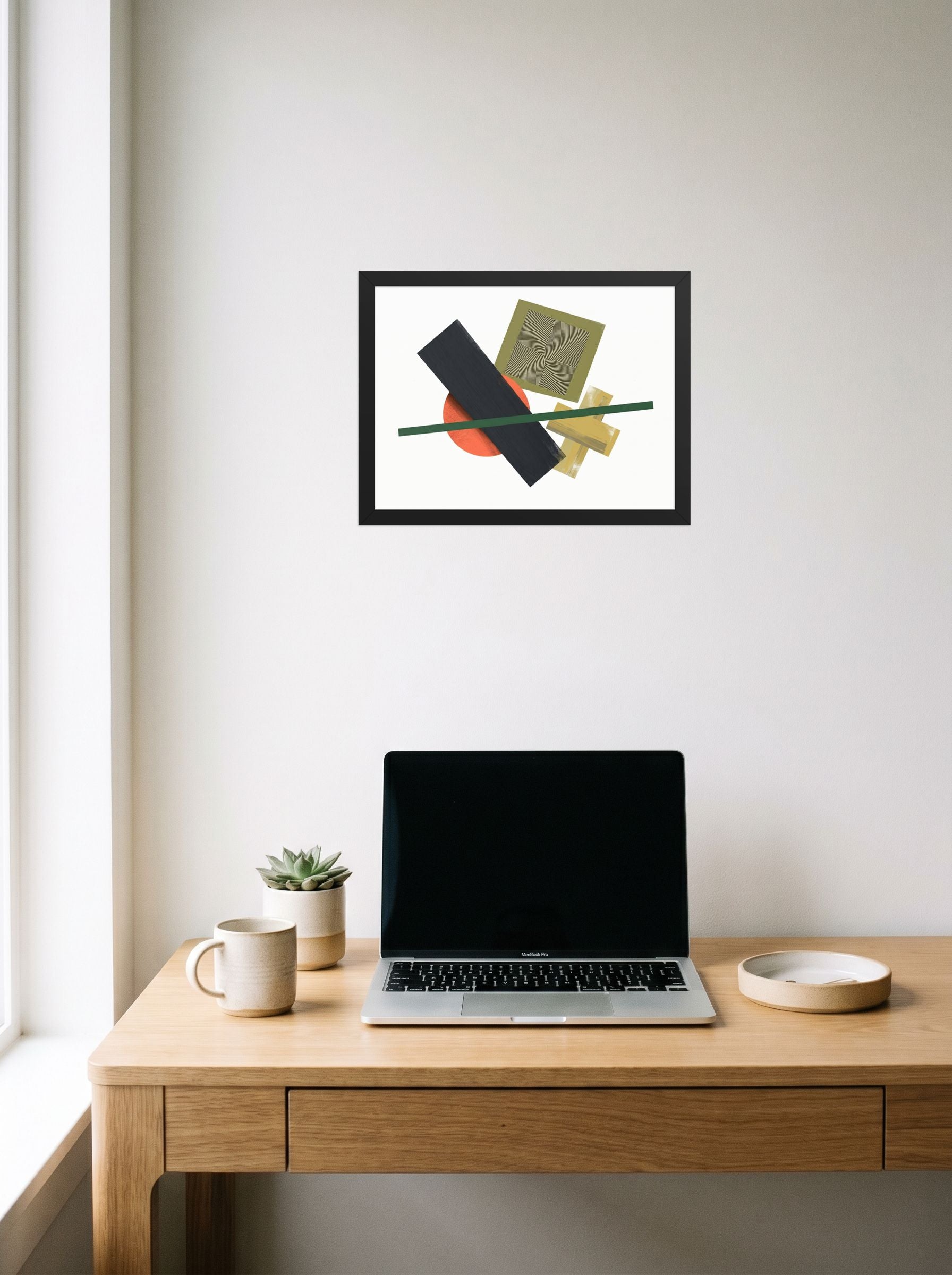 A 16x12 inch black framed Suprematism print of 'The Geometry of Sound' hanging above a worktable with a laptop.