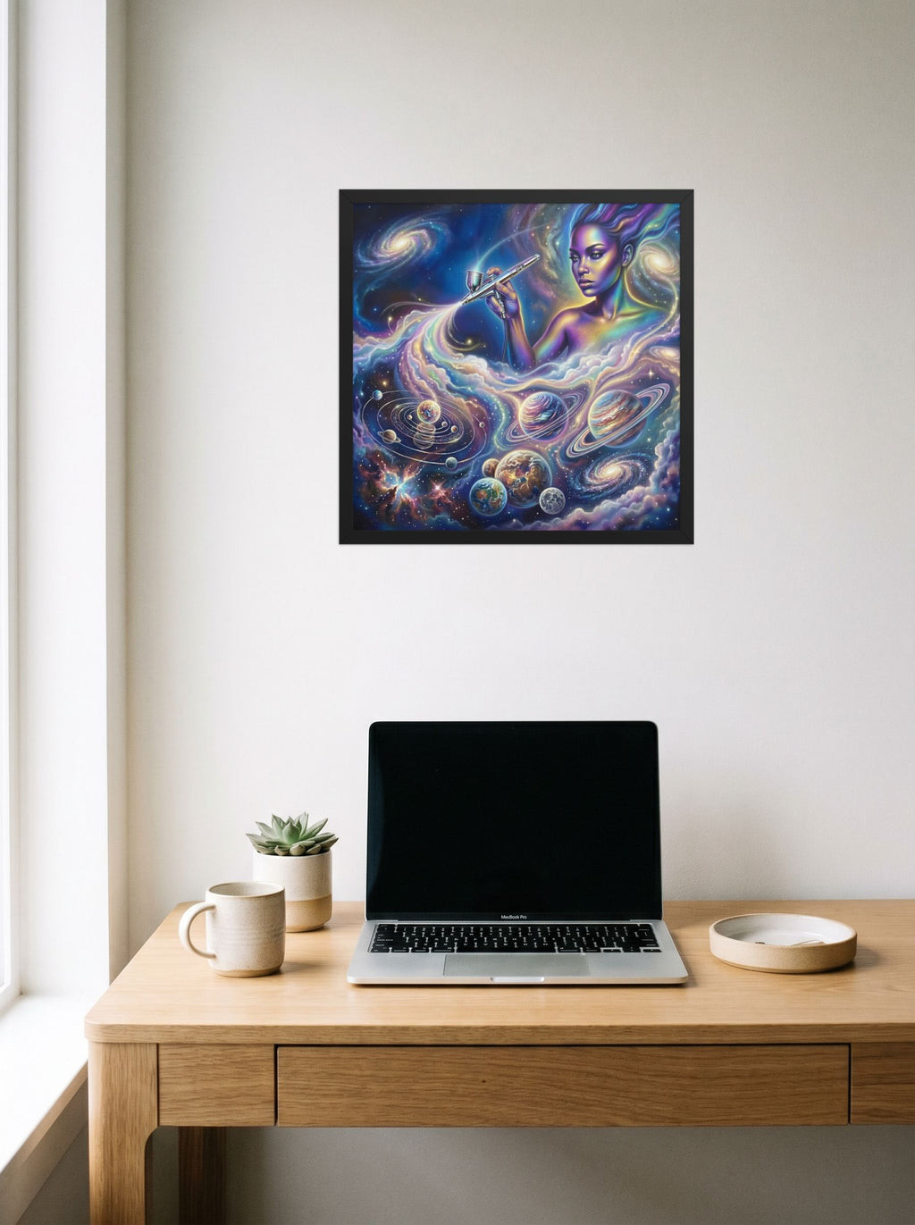 A 18x18 inch black framed Space art print of 'The Cosmic Painter' hanging above a worktable with a laptop.