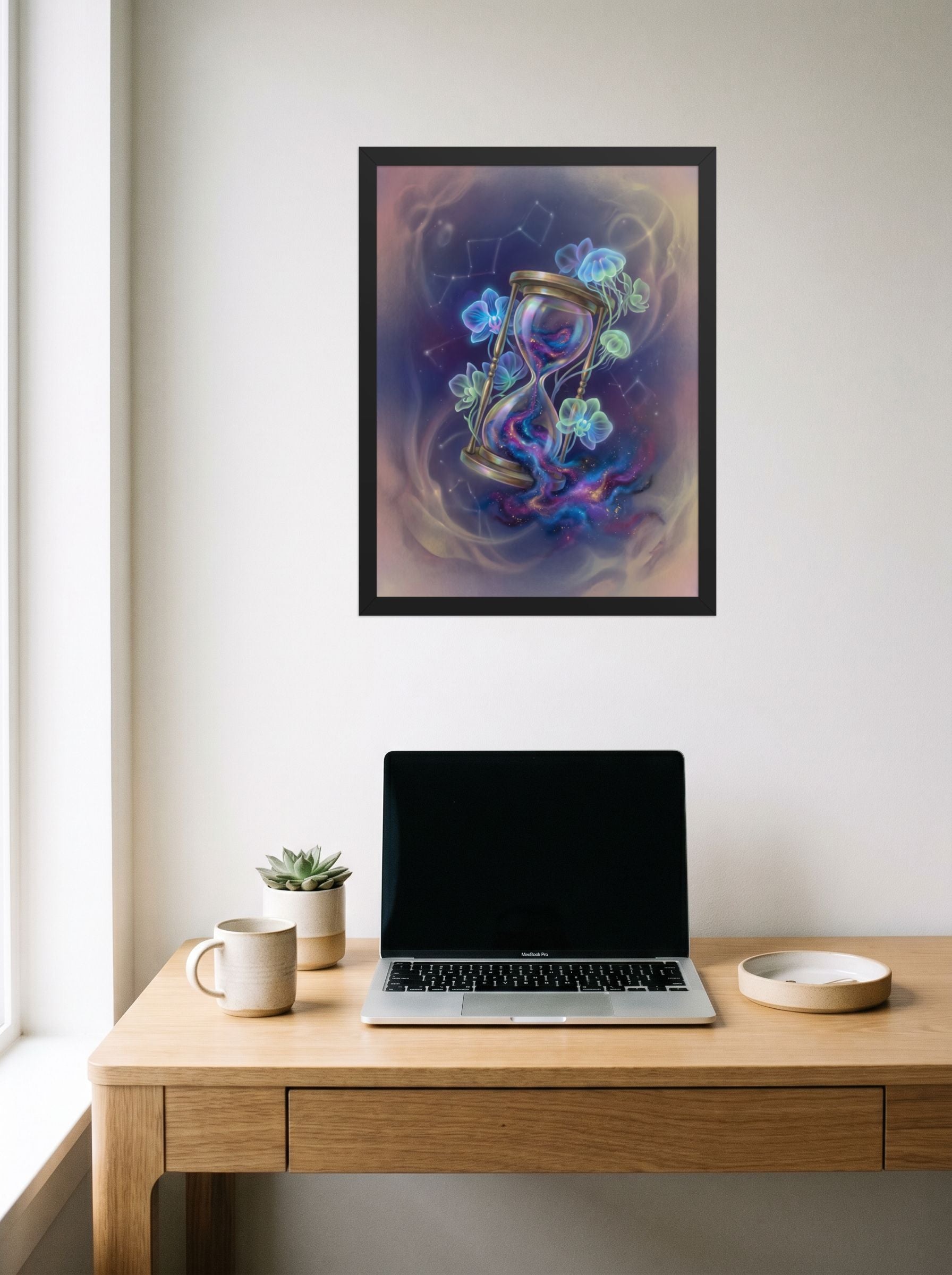 A 18x24 inch black framed Magic realism print of 'Sands of the Nebula' hanging above a worktable with a laptop.