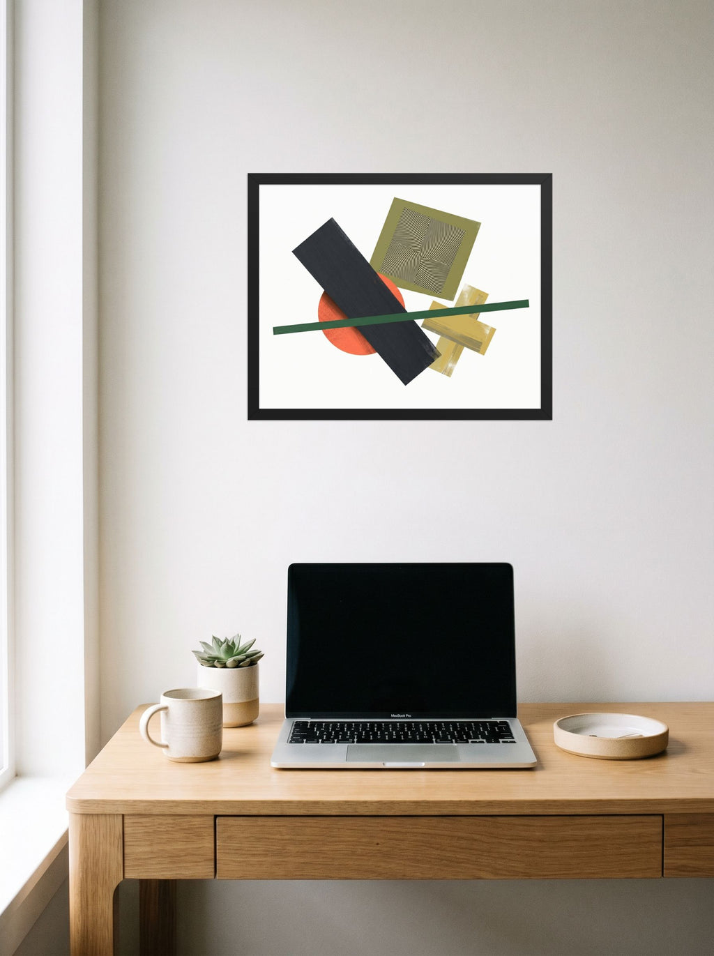 A 20x16 inch black framed Suprematism print of 'The Geometry of Sound' hanging above a worktable with a laptop.