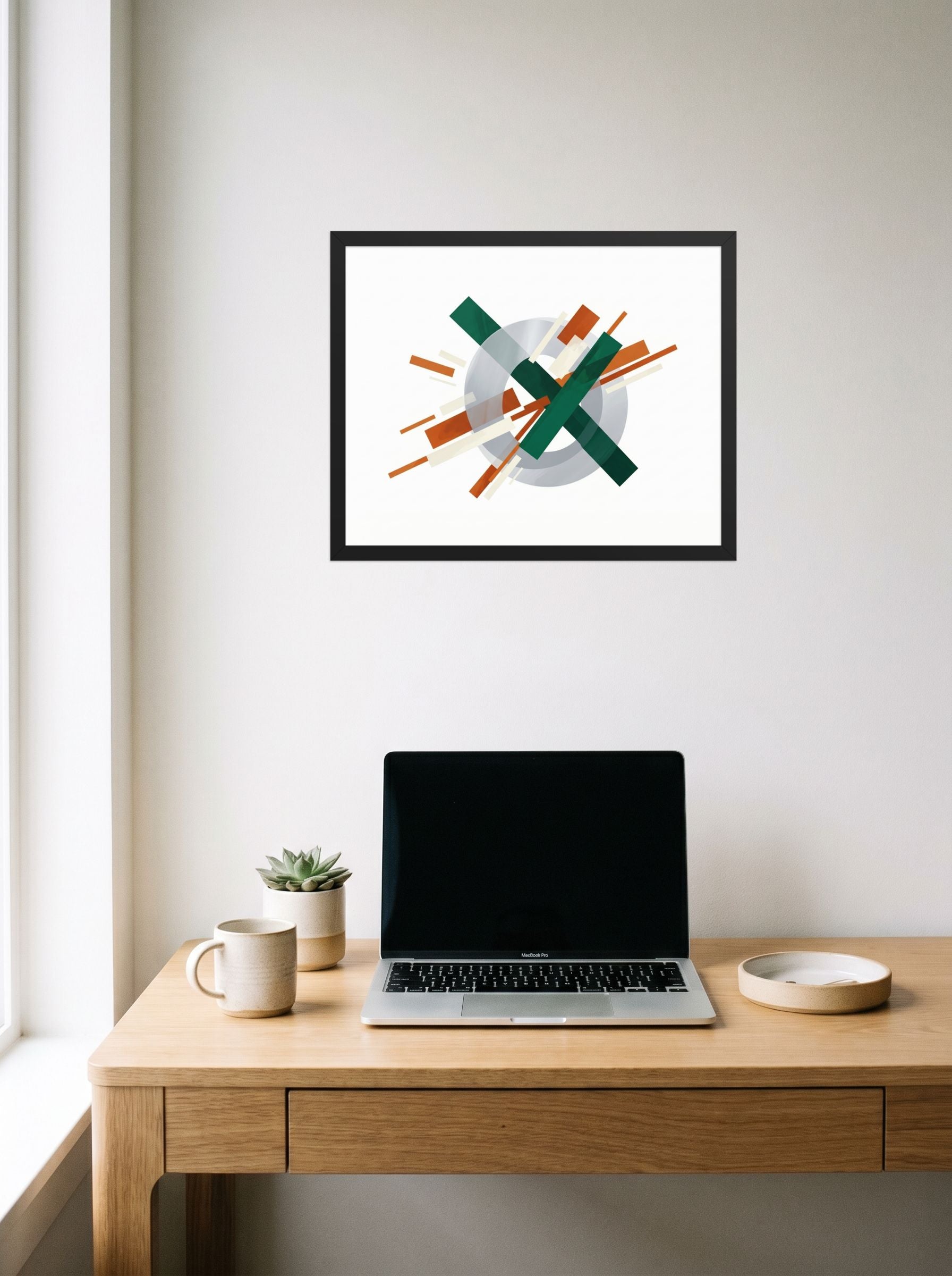 A 20x16 inch black framed Suprematism print of 'Intersecting Perspectives' hanging above a worktable with a laptop.
