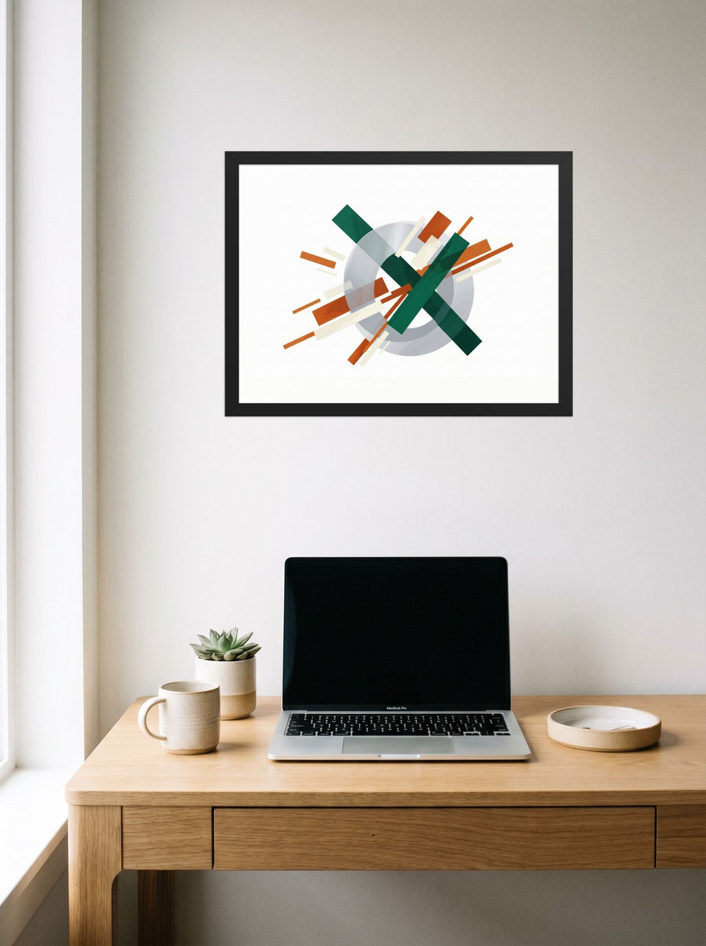 A 24x18 inch black framed Suprematism print of 'Intersecting Perspectives' hanging above a worktable with a laptop.