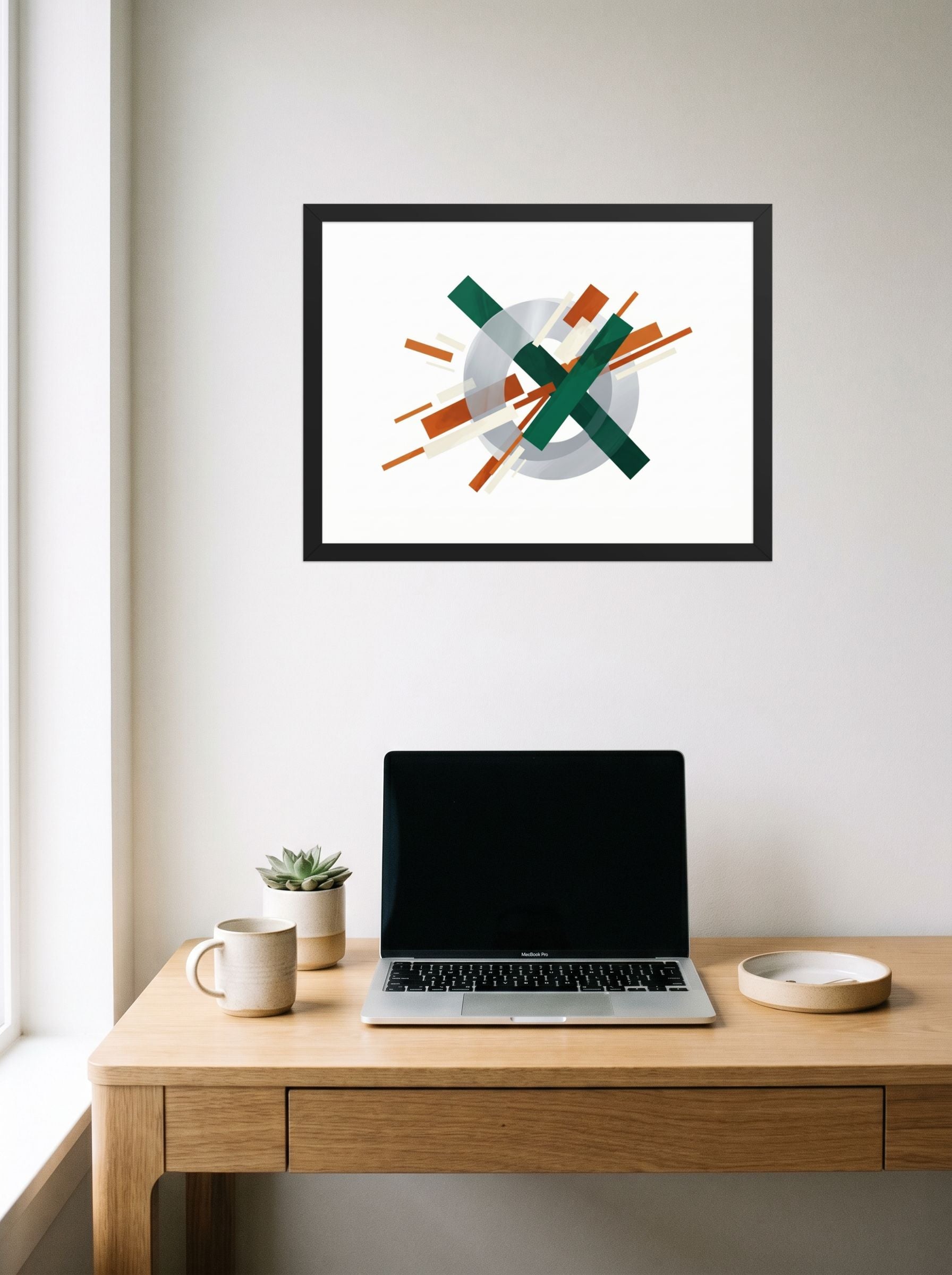 A 24x18 inch black framed Suprematism print of 'Intersecting Perspectives' hanging above a worktable with a laptop.
