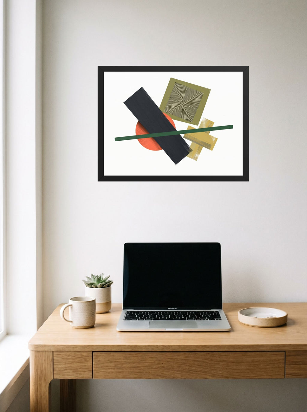 A 24x18 inch black framed Suprematism print of 'The Geometry of Sound' hanging above a worktable with a laptop.