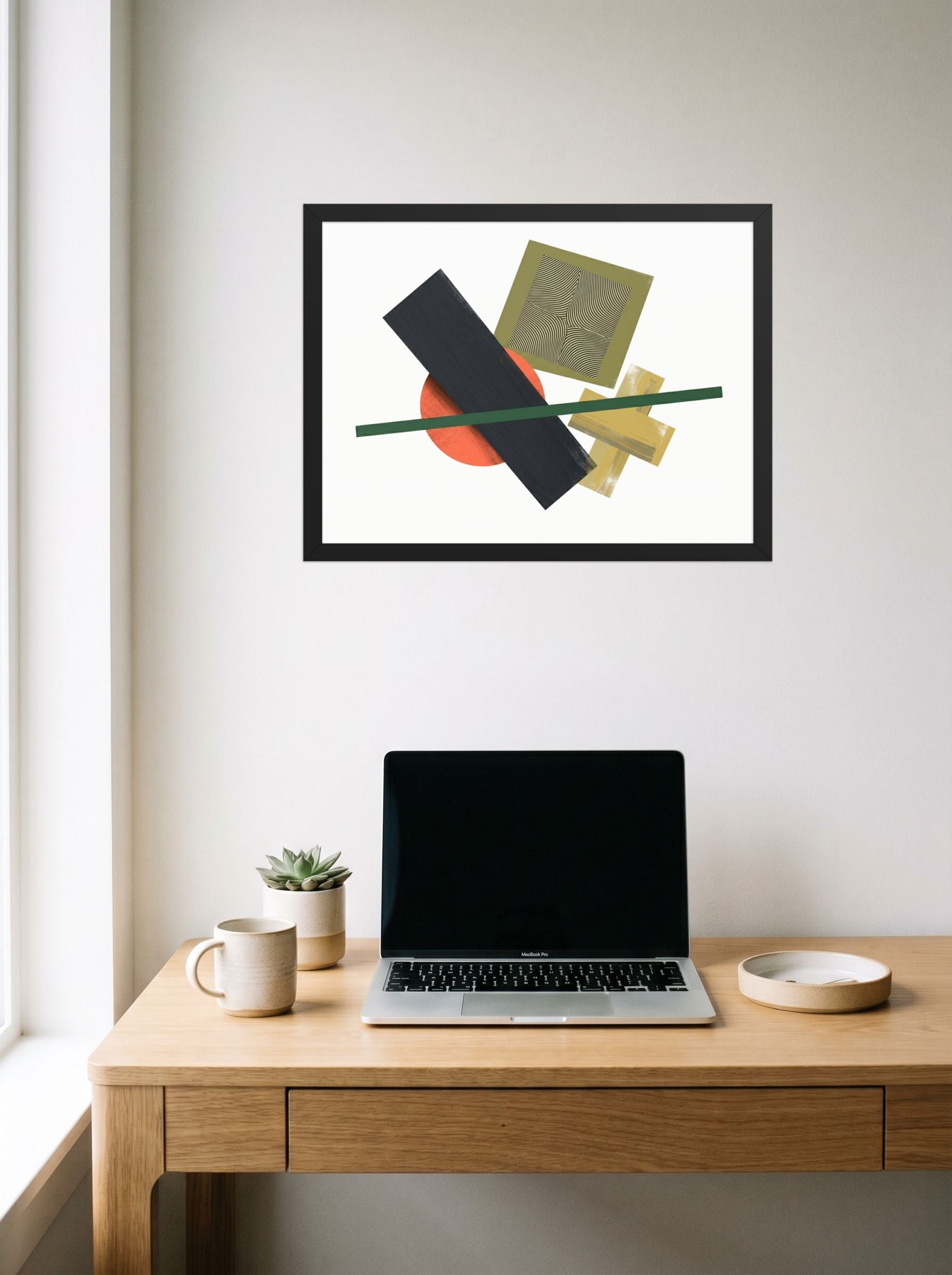 A 24x18 inch black framed Suprematism print of 'The Geometry of Sound' hanging above a worktable with a laptop.