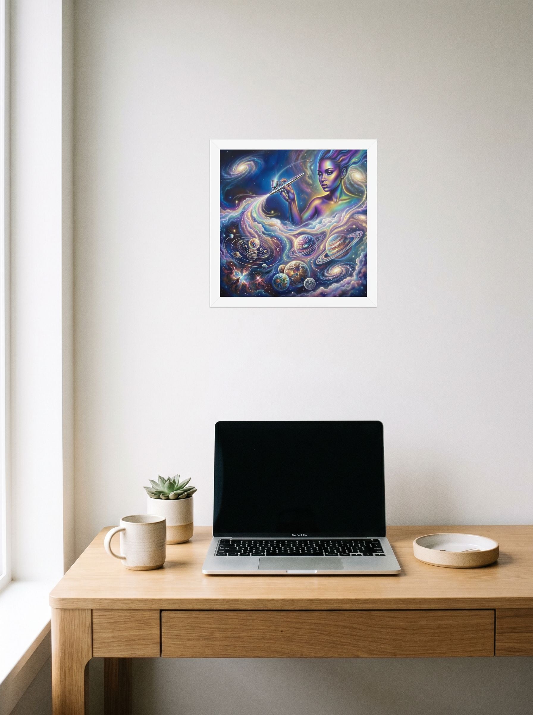 A 12x12 inch white framed Space art print of 'The Cosmic Painter' hanging above a worktable with a laptop.