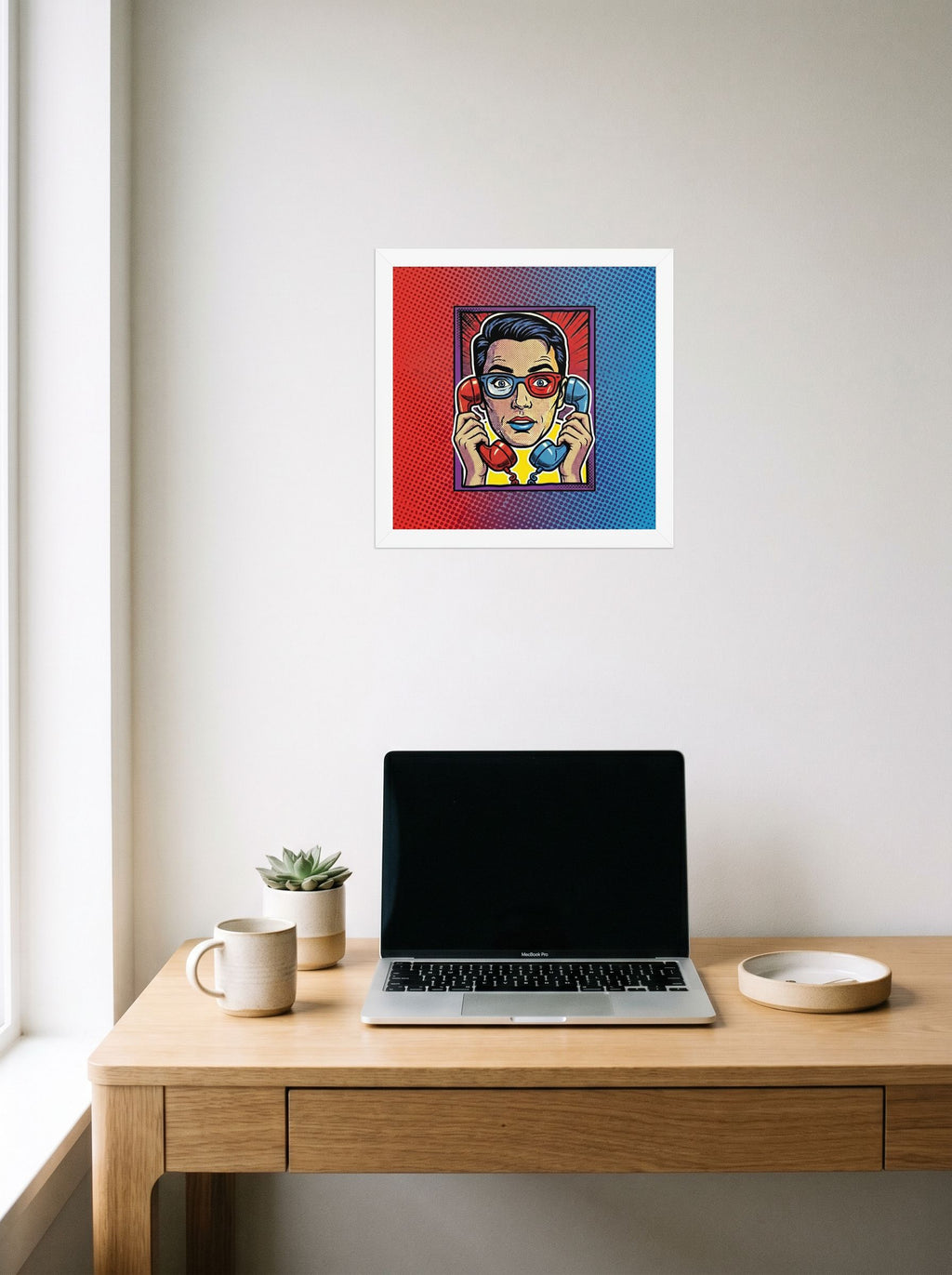A 12x12 inch white framed Pop art print of 'Bipartisan Static' hanging above a worktable with a laptop.