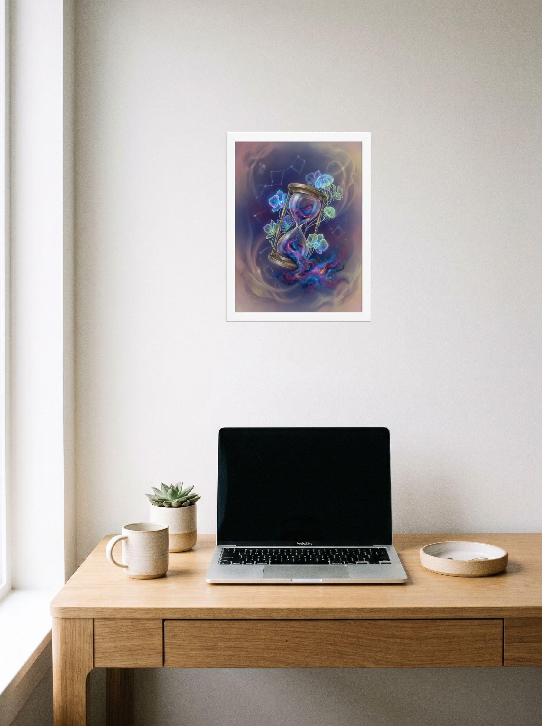 A 12x16 inch white framed Magic realism print of 'Sands of the Nebula' hanging above a worktable with a laptop.