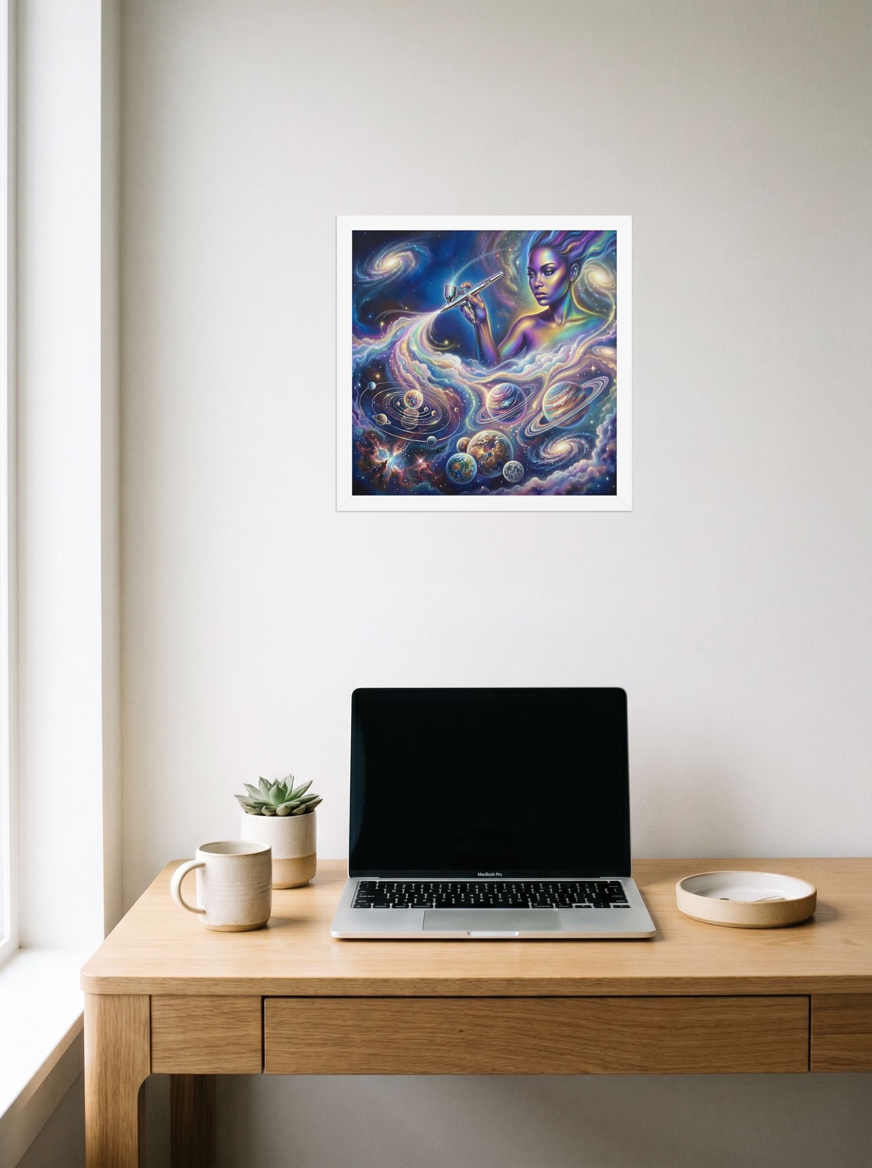 A 14x14 inch white framed Space art print of 'The Cosmic Painter' hanging above a worktable with a laptop.