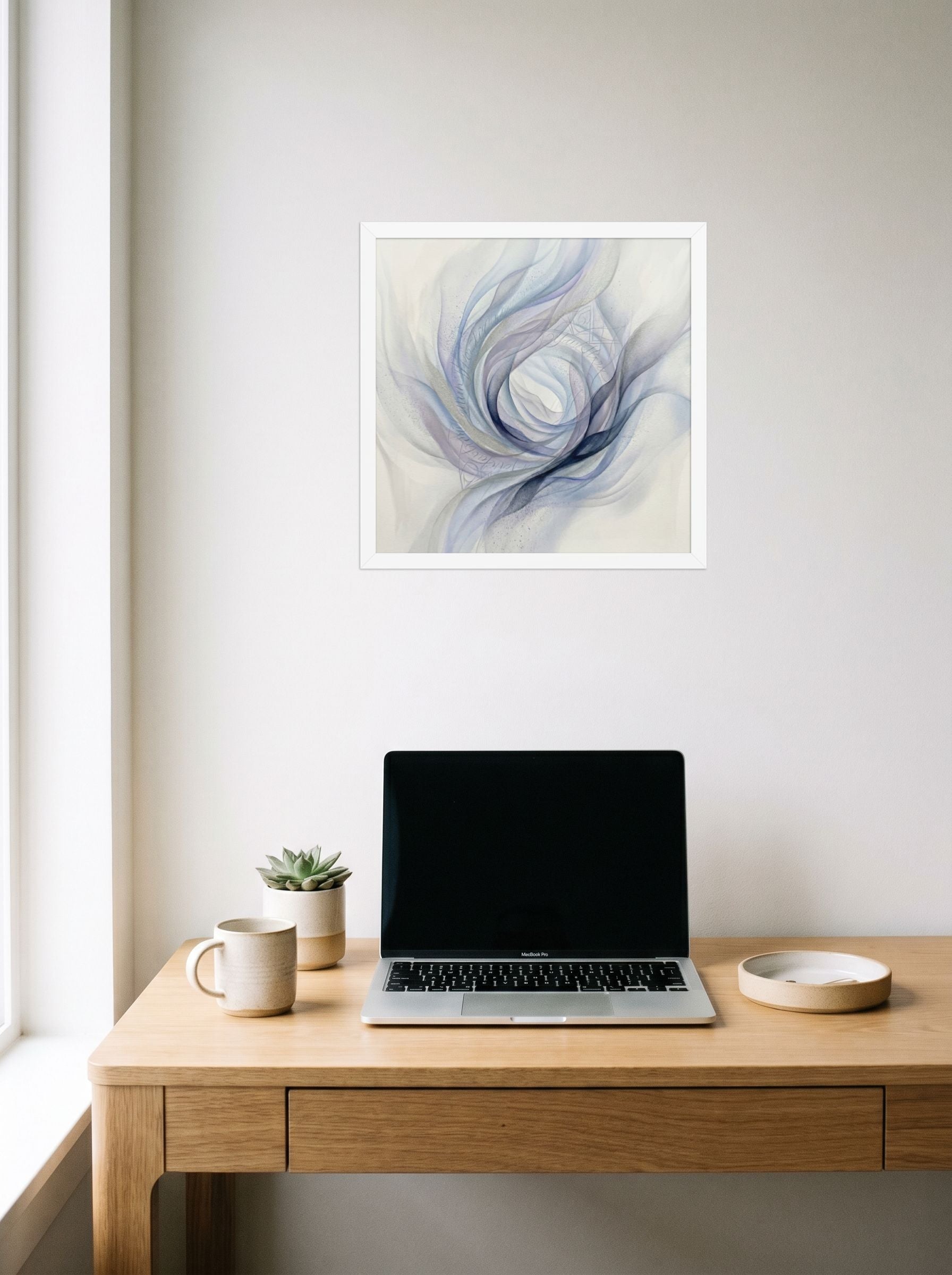 A 16x16 inch white framed Abstract art print of 'Ethereal Whispers in the Vortex' hanging above a worktable with a laptop.