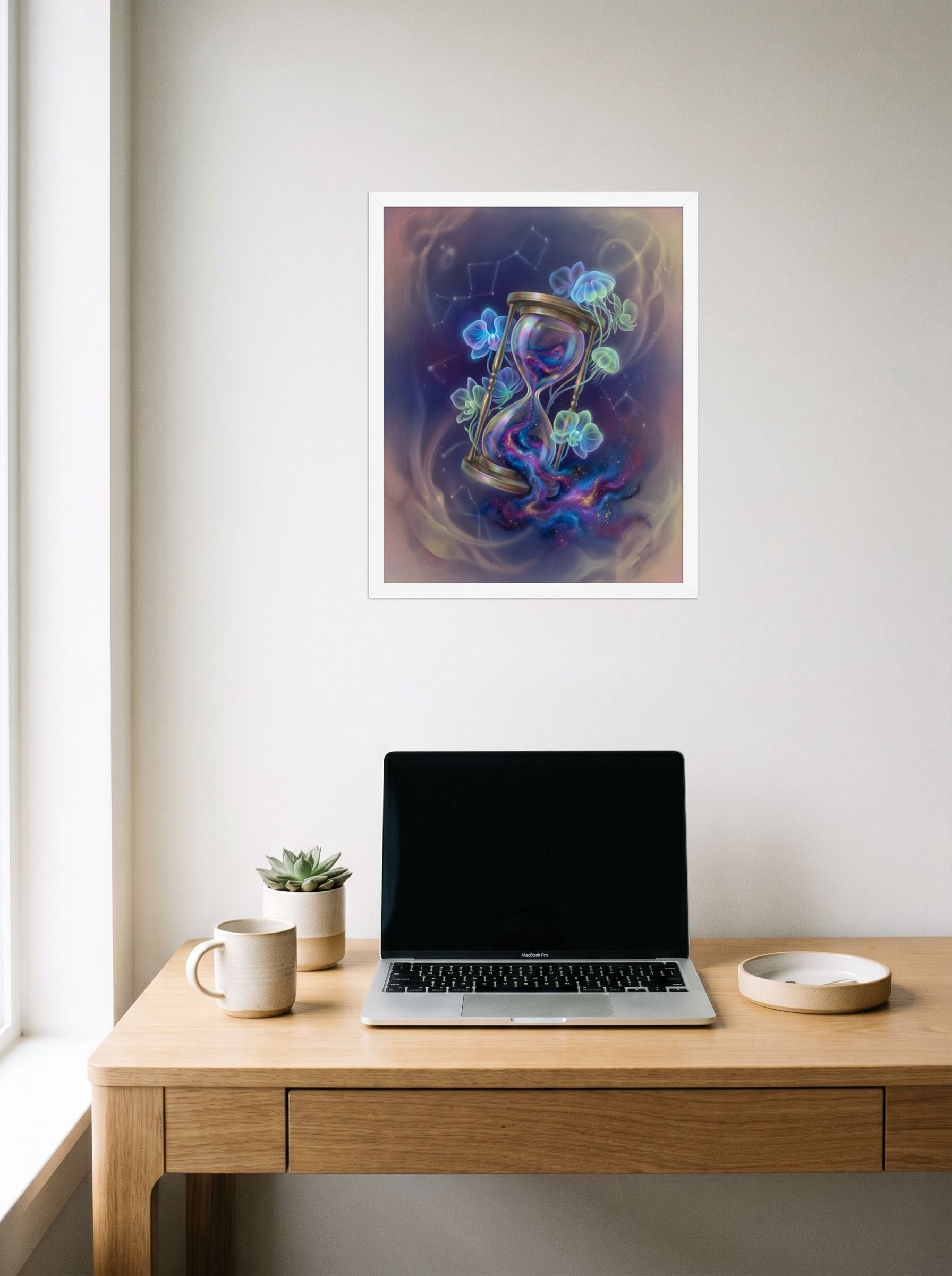 A 16x20 inch white framed Magic realism print of 'Sands of the Nebula' hanging above a worktable with a laptop.