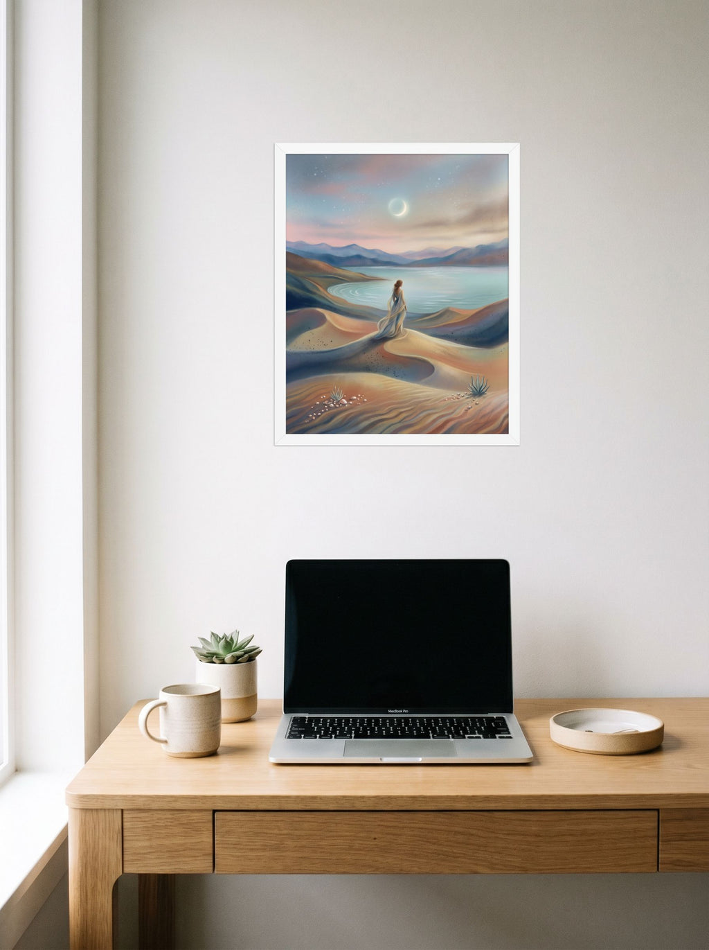 A 16x20 inch white framed Magic realism print of 'Desert Moonrise' hanging above a worktable with a laptop.