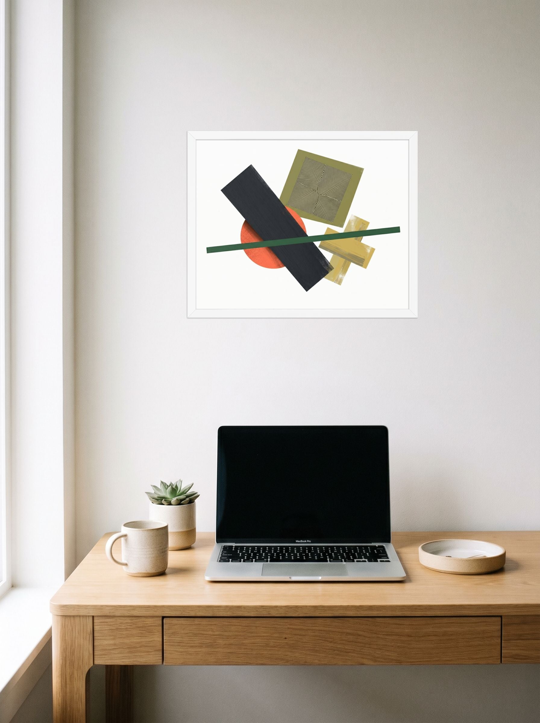 A 20x16 inch white framed Suprematism print of 'The Geometry of Sound' hanging above a worktable with a laptop.