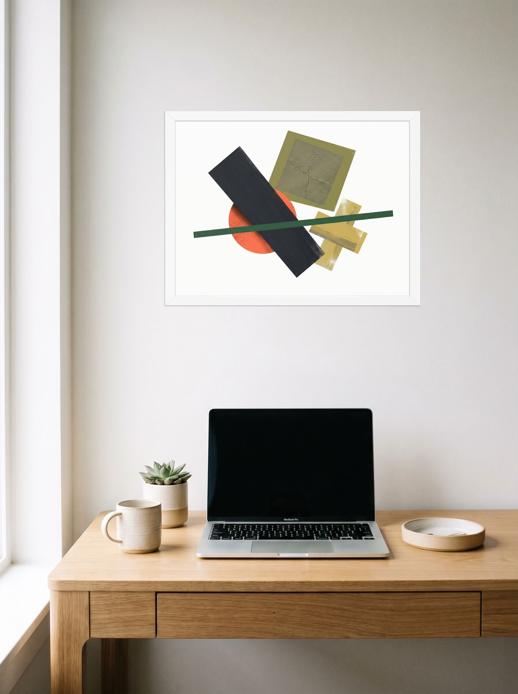 A 24x18 inch white framed Suprematism print of 'The Geometry of Sound' hanging above a worktable with a laptop.
