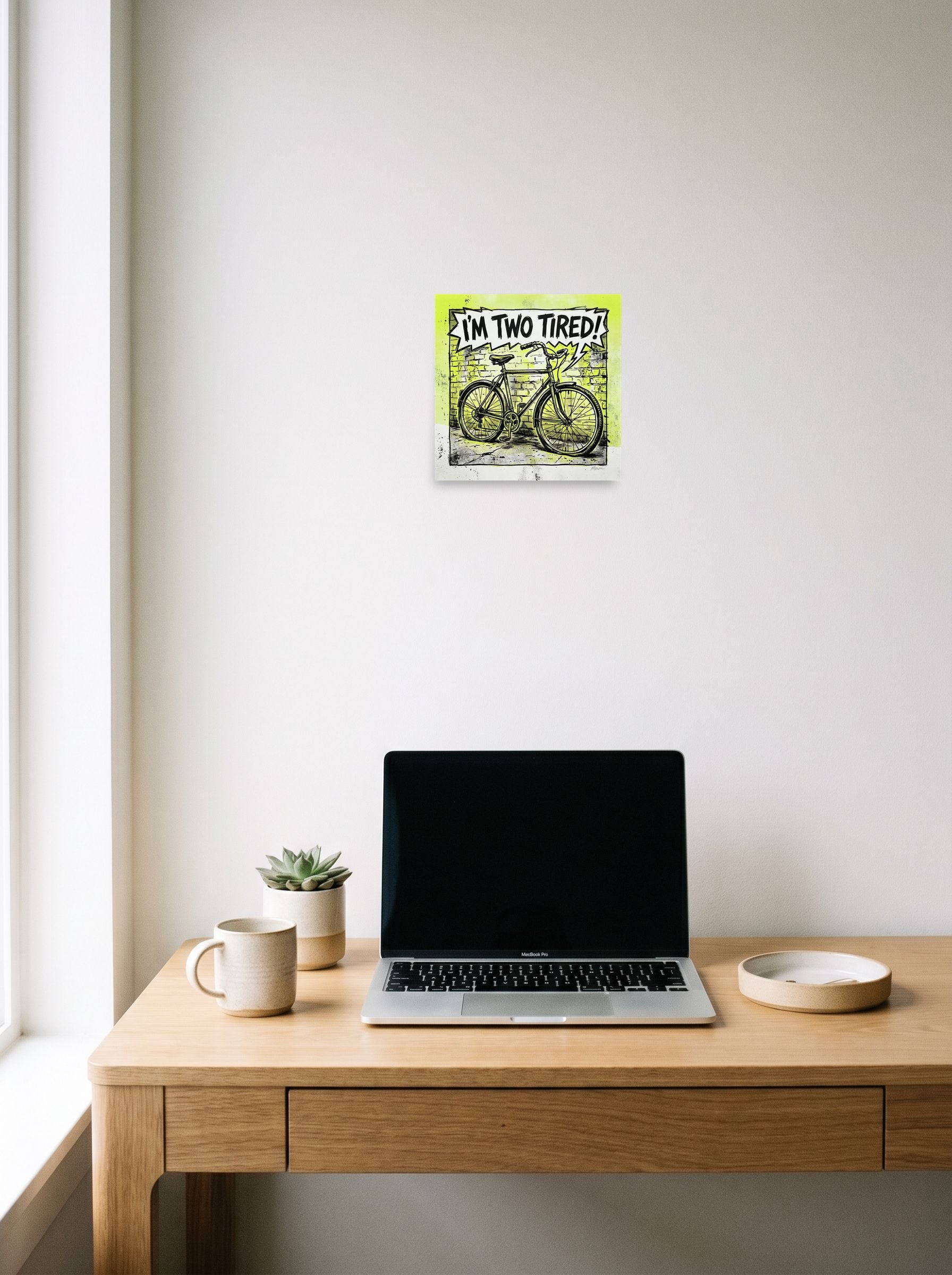 A 10x10 inch unframed paper Pop art print of 'Two Tired' hanging above a worktable with a laptop.