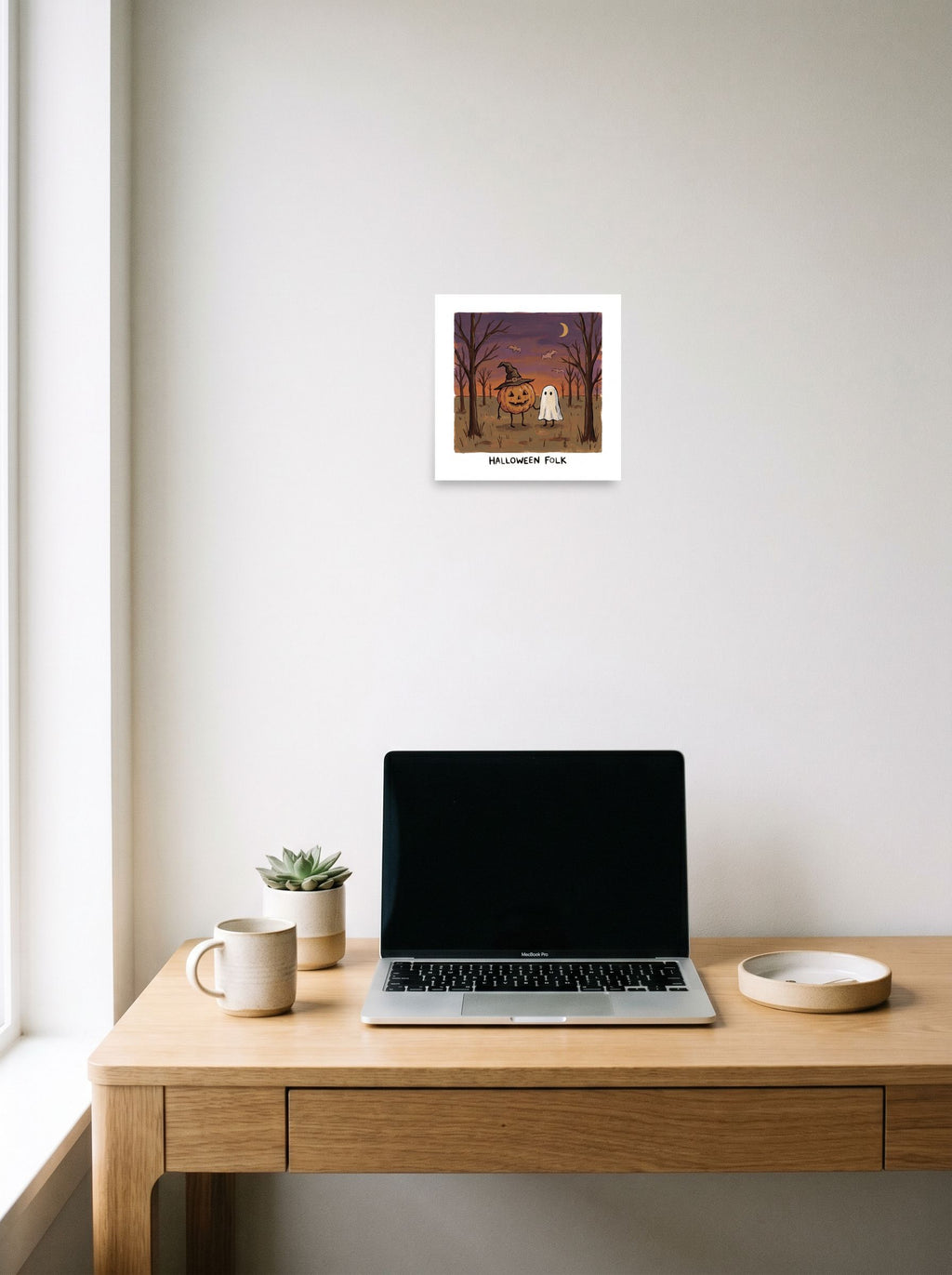 A 10x10 inch unframed paper Naive art print of 'Halloween Folk' hanging above a worktable with a laptop.