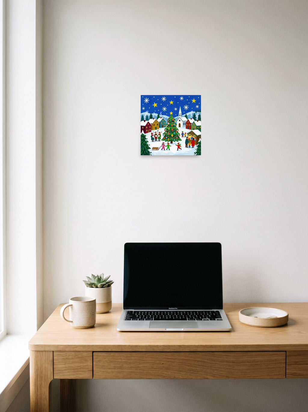 A 10x10 inch unframed paper Naive art print of 'Joyous Christmas Village' hanging above a worktable with a laptop.