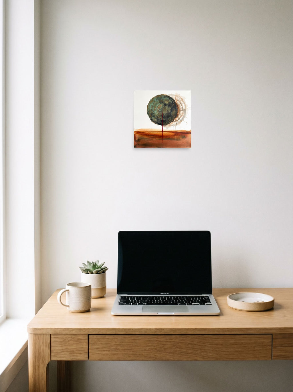 Mockup of 'Orb of Verdigris' as a 10x10 inch unframed paper print, hanging above a worktable with a laptop.