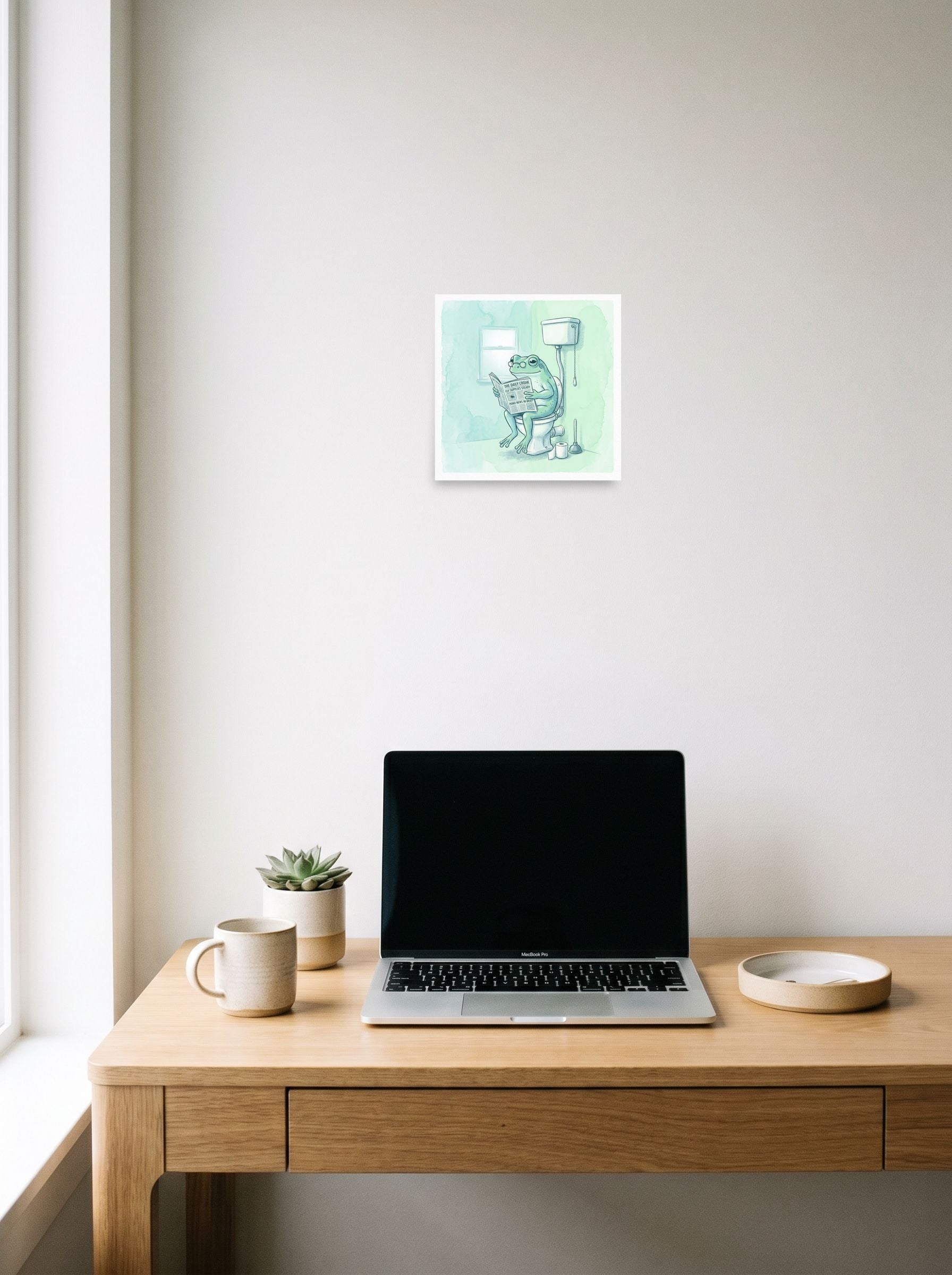A 10x10 inch unframed paper Realism print of 'The Daily Croak' hanging above a worktable with a laptop.