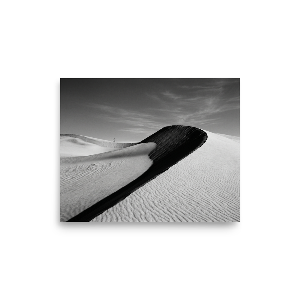 'Footsteps in the Void' — a Art photography print. A minimalist black and white photograph of sweeping sand dunes with a tiny figure walking along the ridge, perfect for adding a sense of scale to an Office.