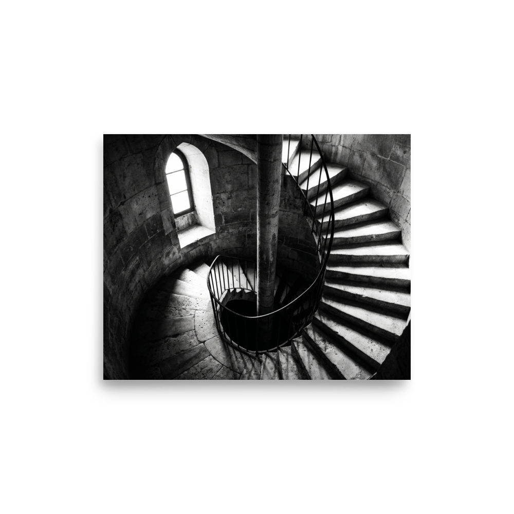 'Descent into Light' — a Art photography print. A high-contrast black and white photograph looking down a spiral stone staircase illuminated by a small window, adding depth and intrigue to an Office or Hallway.