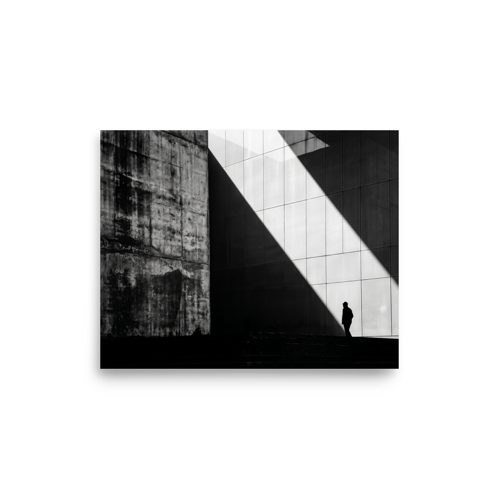 'Urban Geometry' — a Modernism print. A high-contrast black and white architectural photograph featuring a sharp diagonal shaft of light and a lone silhouette walking past textured concrete walls, perfect for an Office or Modern Living Room.