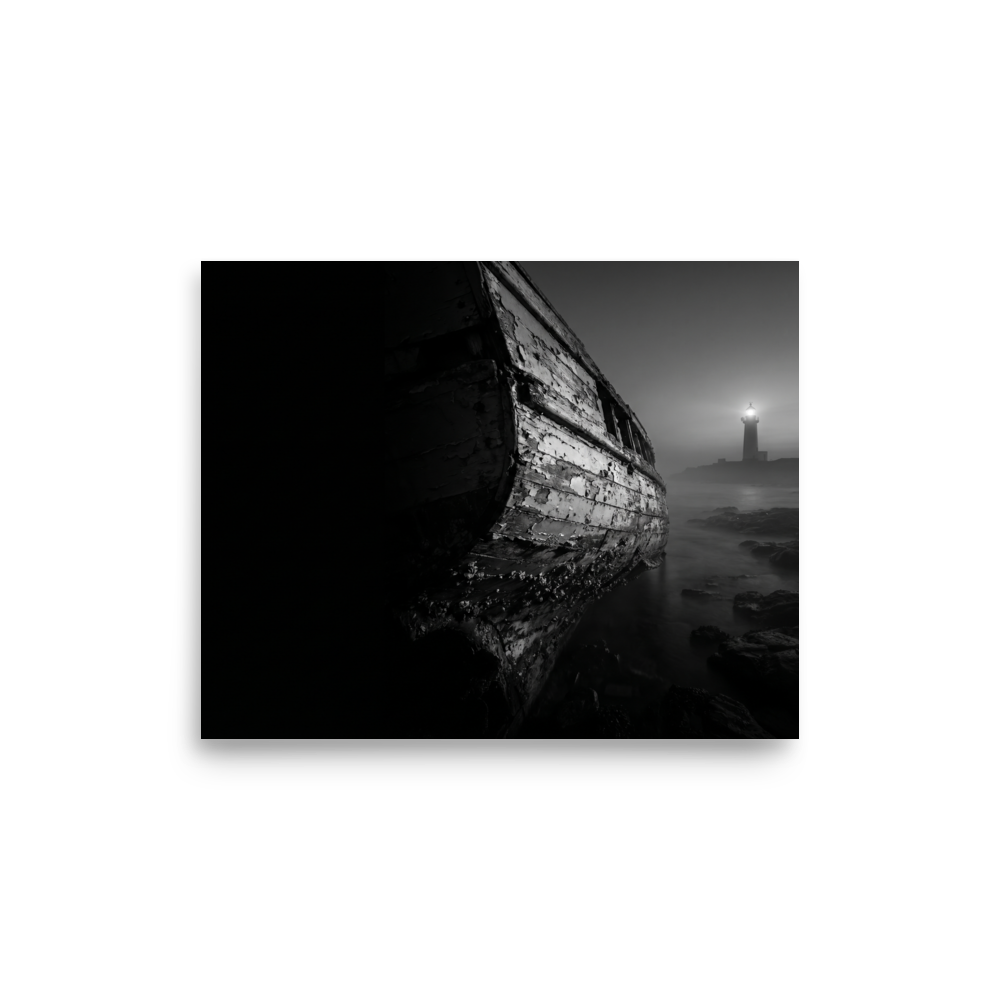 'Final Anchor' — a Art photography print. Black and white photograph of an old, peeling wooden boat resting on rocks with a glowing lighthouse in the foggy distance, perfect for a cozy living room.