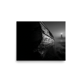 'Final Anchor' — a Art photography print. Black and white photograph of an old, peeling wooden boat resting on rocks with a glowing lighthouse in the foggy distance, perfect for a cozy living room.