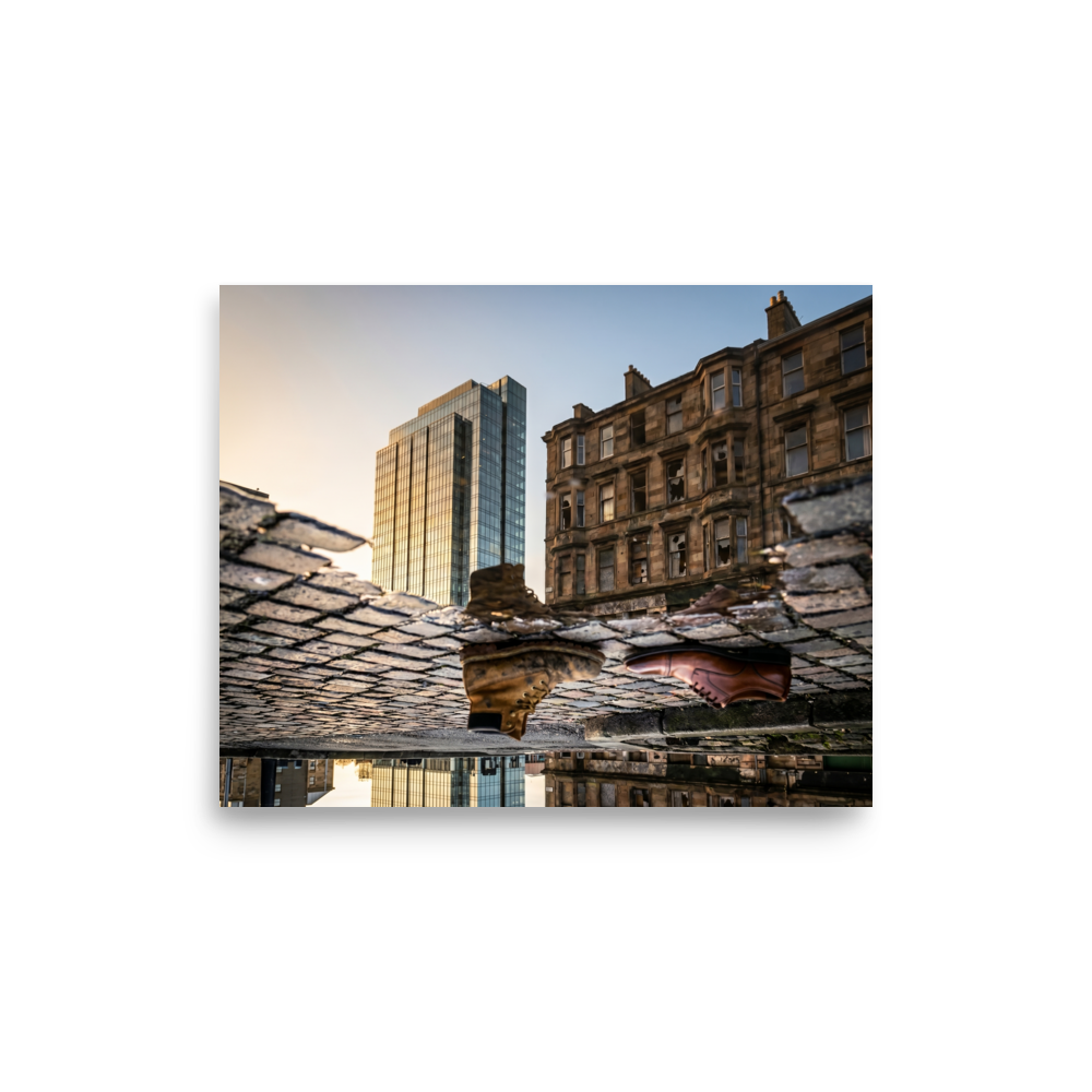 'Steps Through Time' — a Art photography print. Creative puddle reflection photograph showing a work boot and a dress shoe on cobblestones, with a modern glass skyscraper and an old stone building in the background, ideal for an office or hallway.