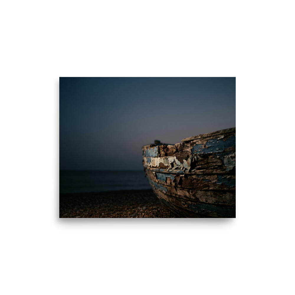 'The Faded Blue Boat' — a Art photography print. Close-up photograph of a weathered wooden boat with peeling blue paint resting on a rocky beach at dusk, suitable for a serene bathroom or living room.